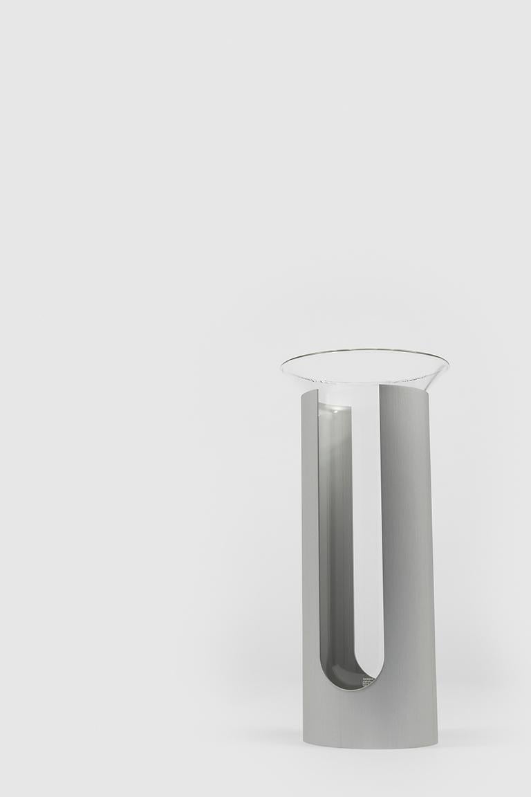 Camicia is a vase made up of two elements: a cylinder of opaque anodized aluminium without a bottom that supports and envelops a transparent glass container. The round hole on one side of the metal cylinder and the opening that runs vertically from