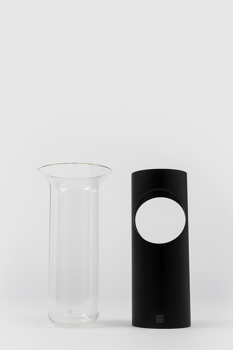 Camicia is a vase made up of two elements: a cylinder of opaque anodized aluminium without a bottom that supports and envelops a transparent glass container. The round hole on one side of the metal cylinder and the opening that runs vertically from