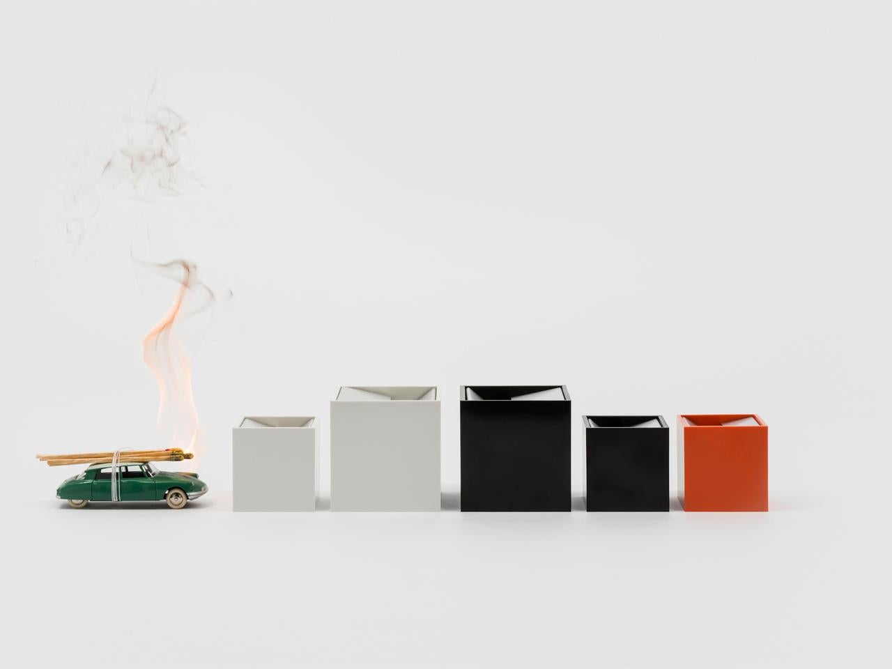 Larger White Cubo is an ashtray made up of two simple elements: a cube shaped shell open on one side contains an element made of bent sheet metal. This element is designed so that the acute angle of one end of the sheet makes the ash fall inside