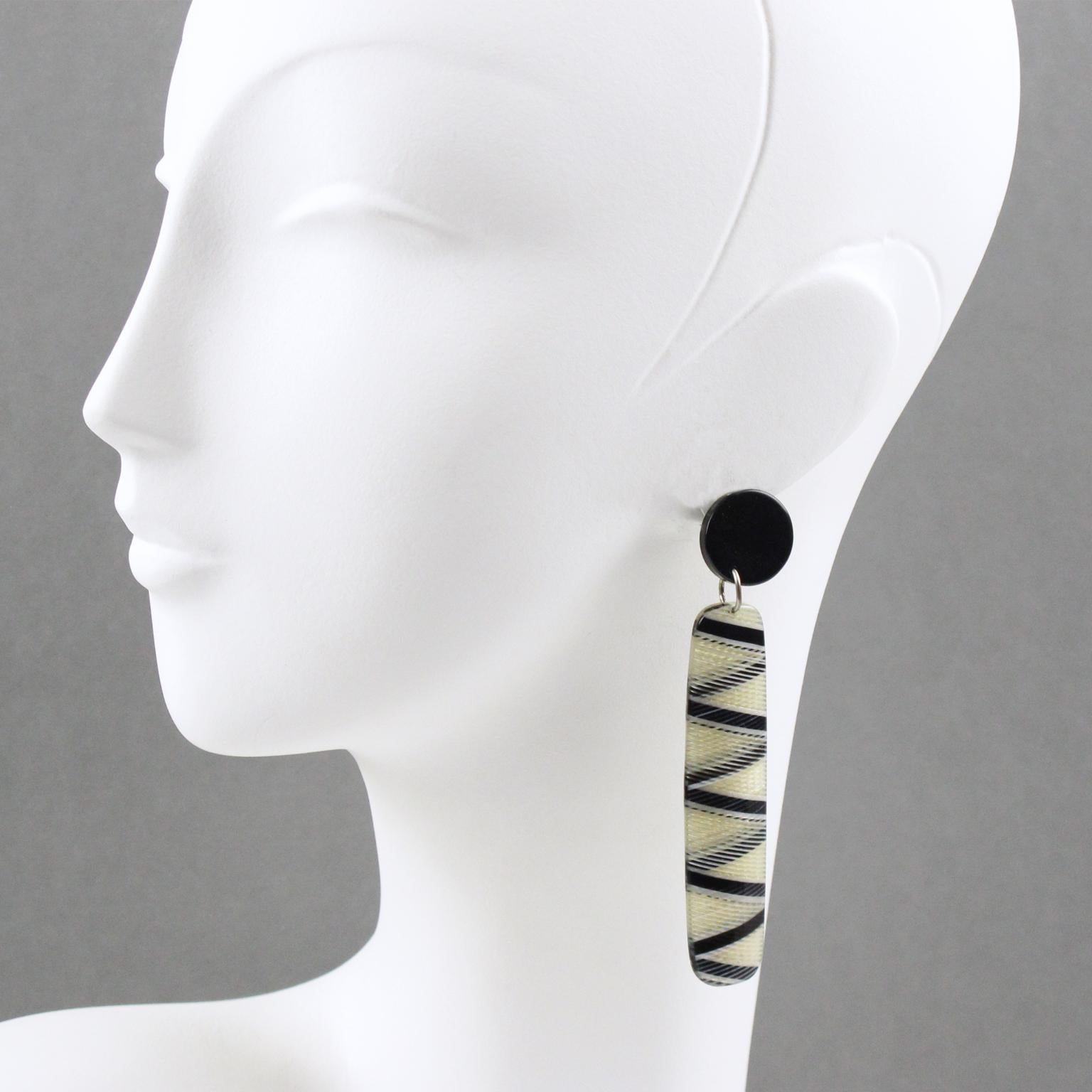 Questi eleganti orecchini pendenti in lucite sono caratterizzati da una lunga forma a goccia con moderne strisce e motivi geometrici. I pezzi sono di colore nero, trasparente e bianco. Non è visibile il marchio del produttore. Questi orecchini sono