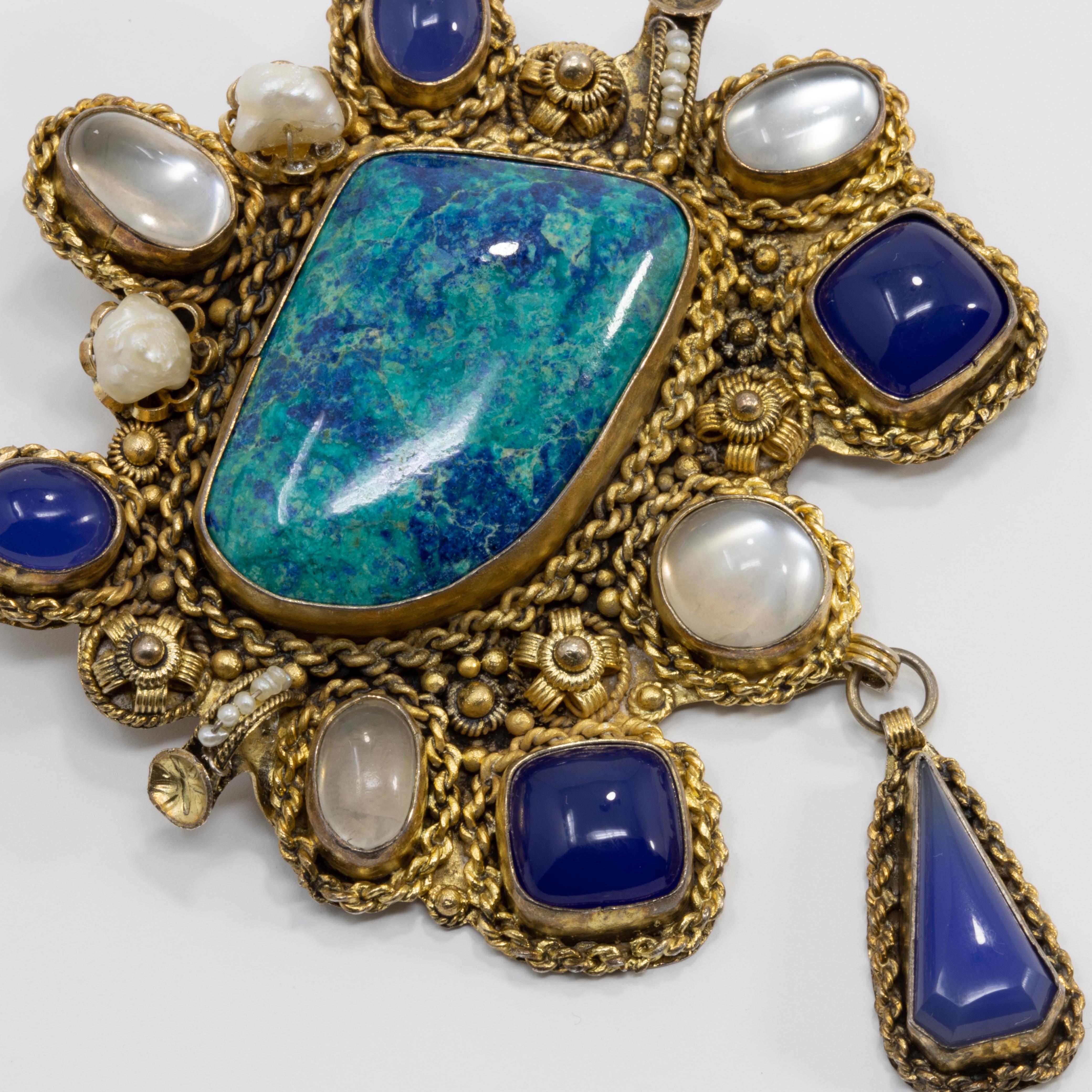 A large pendant with a dangling motif. Perfect when you need to make a statement! Features an assortment of natural gemstones including quartz and mother of pearl. Colors include an assortment of opaque white, blue, and teal. Set in an accented gilt