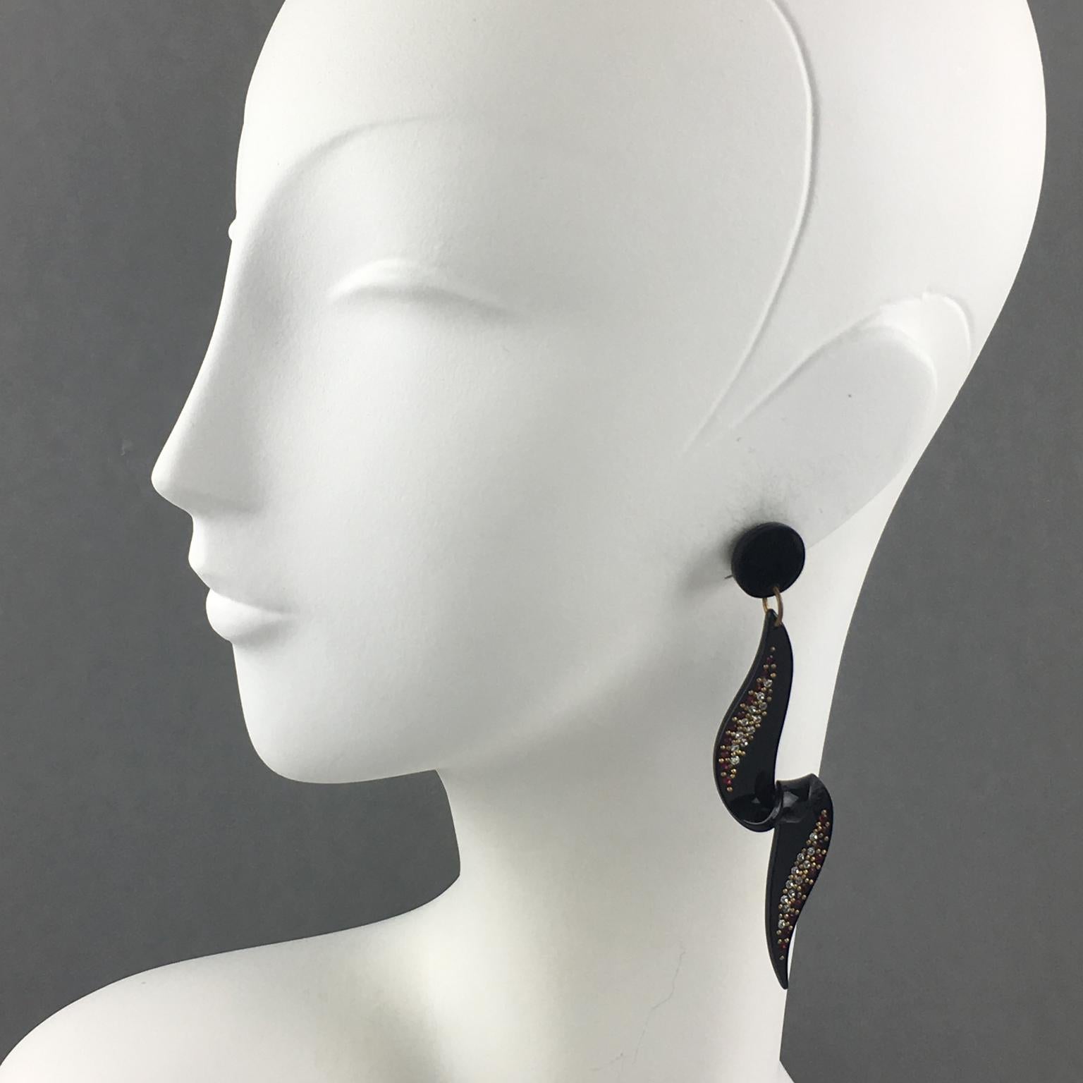 Questi spettacolari orecchini a clip lunghi in lucite o resina presentano un motivo geometrico dimensionale a spirale in colore nero lucido, ornato da piccoli strass rossi e trasparenti e borchie in ottone. Hanno il normale retro a clip francese.