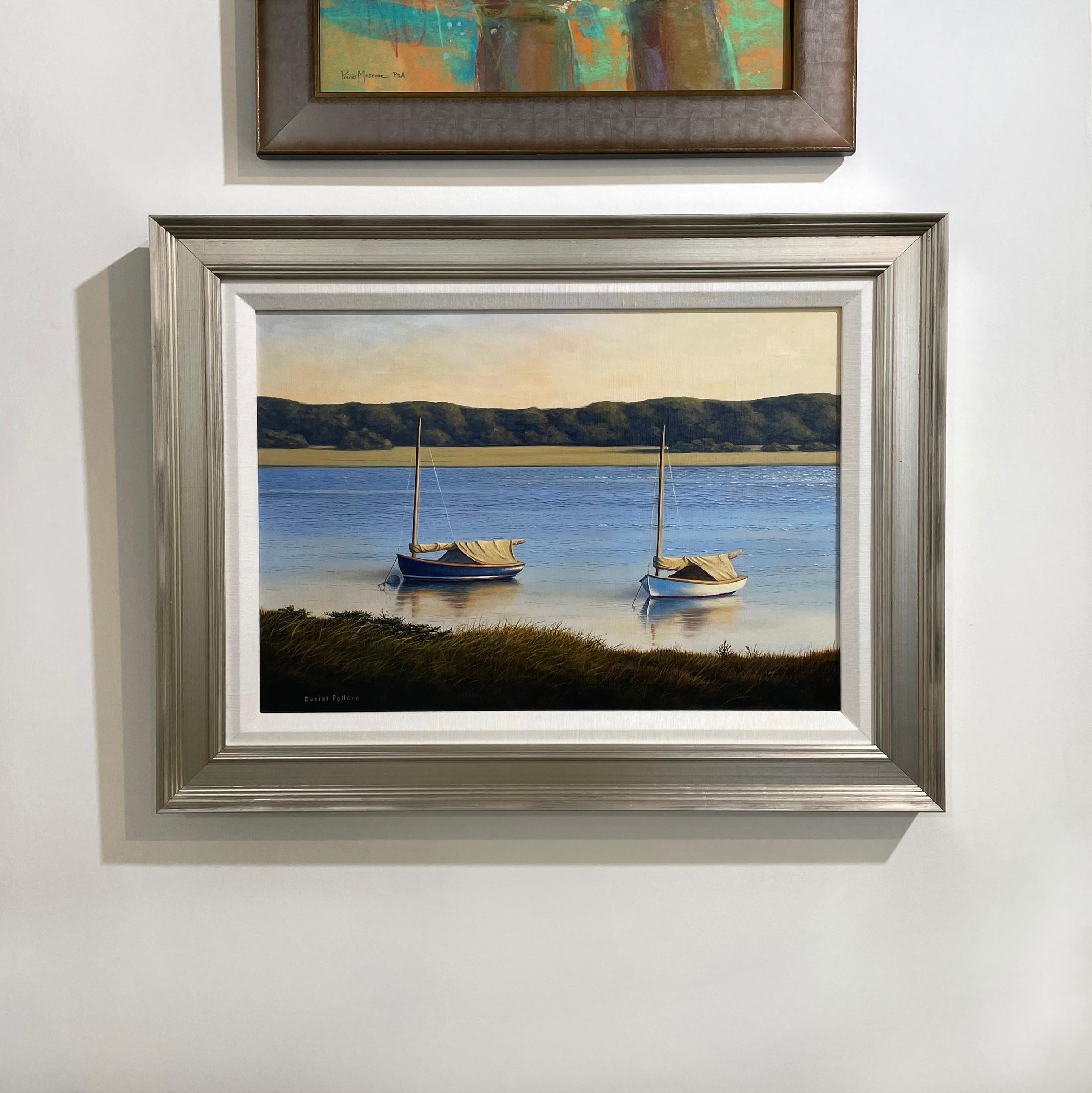 This traditional coastal oil painting by contemporary realist painter Daniel Pollera captures two small sailboats, one white and one navy blue, anchored along a grassy shoreline in the foreground. The boats' reflections are visible in the pale