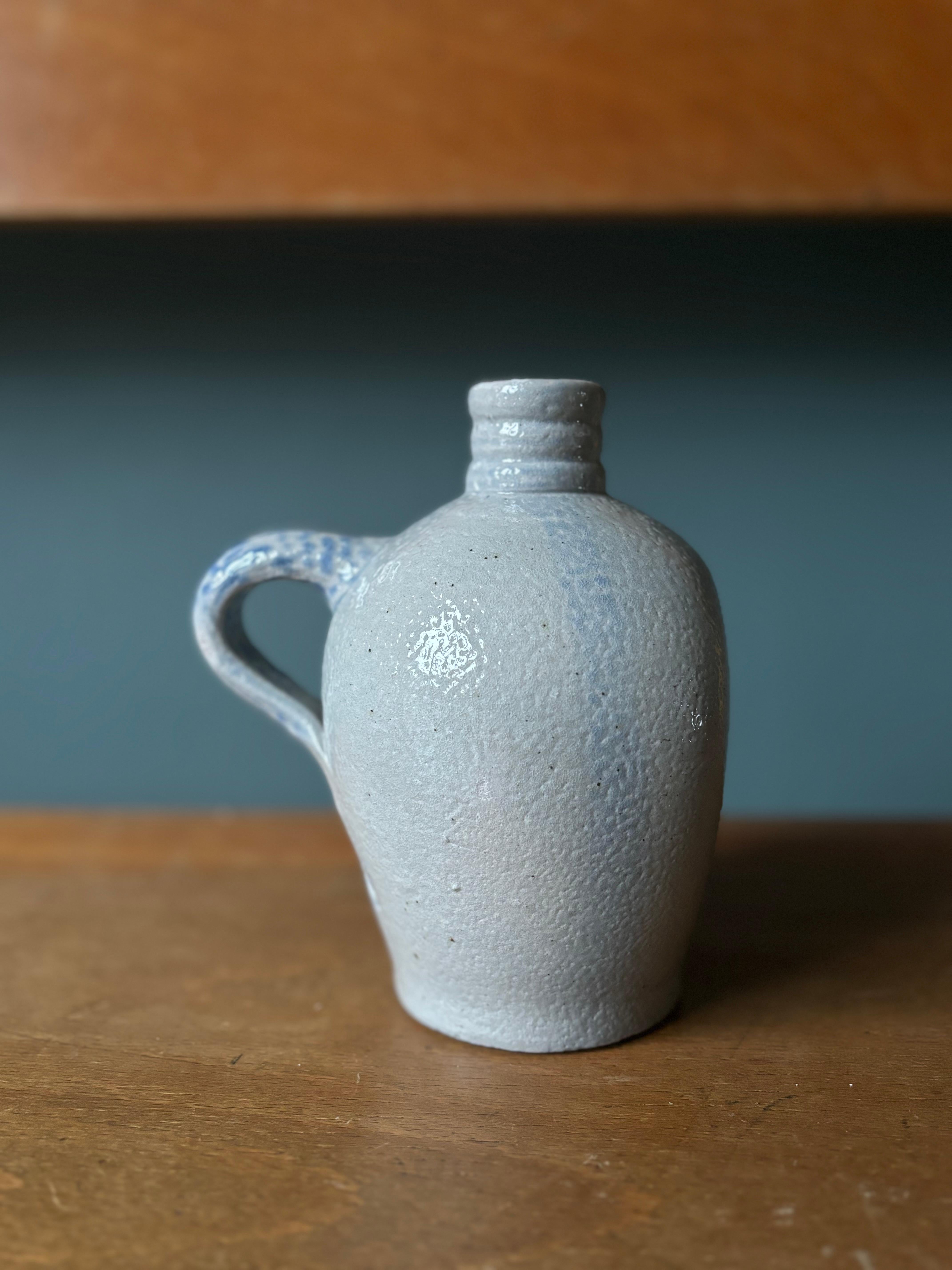 Dekorative Flaschenvase aus Steingut, die ursprünglich in den 1940er Jahren in Dänemark für alkoholische Getränke verwendet wurde. Dicke hellgraue Salzglasur mit babyblauen Flecken entlang des Henkels und der Seiten. Asymmetrische Form mit flacher