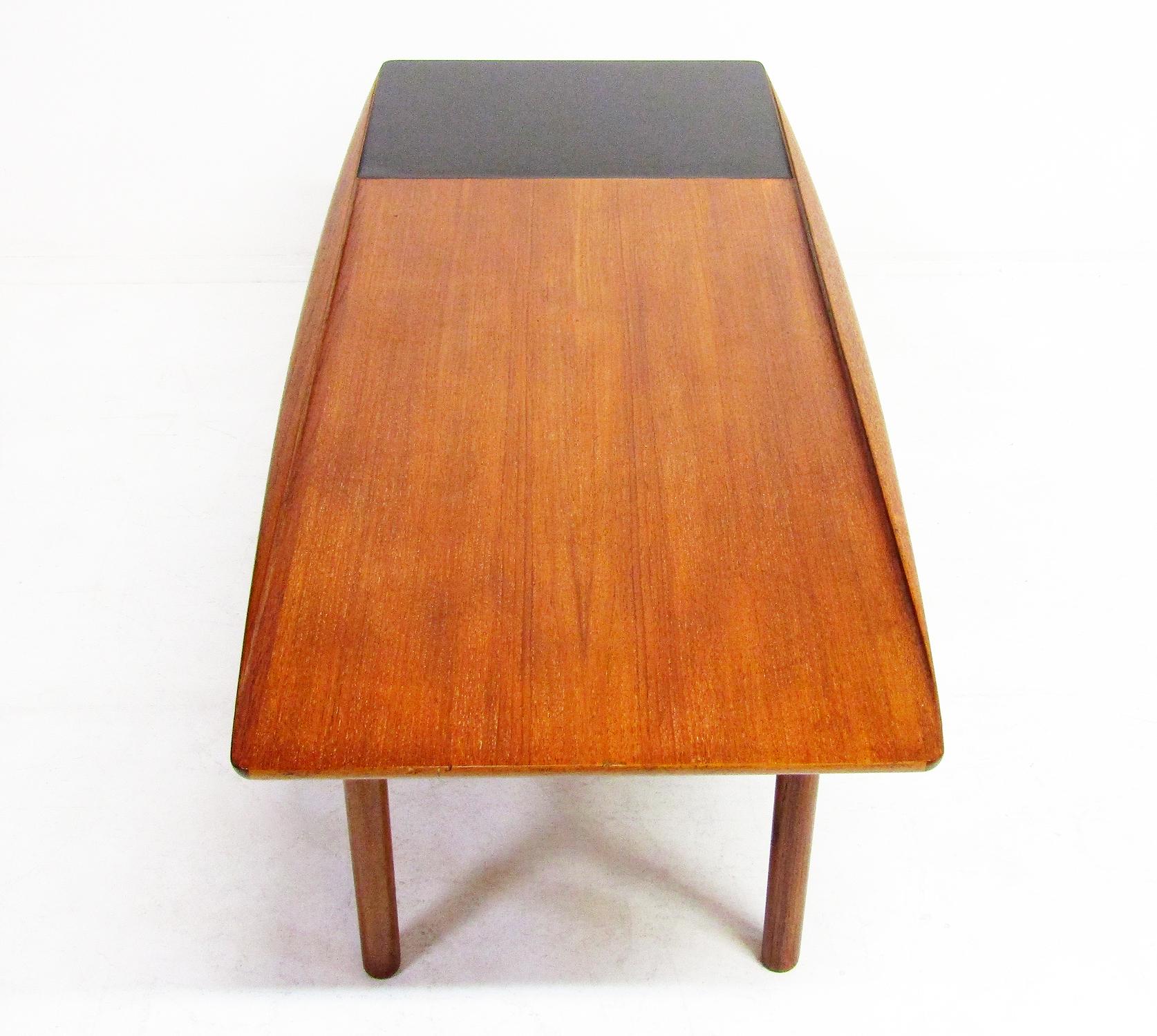A Danish 1950s surfboard coffee table by Grete Jalk for Poul Jeppesen.

It has beautiful sculpted contours. The teak is a rich shade of reddish gold. There is a black formica area for drinks

The Poul Jeppesen label is affixed to the
