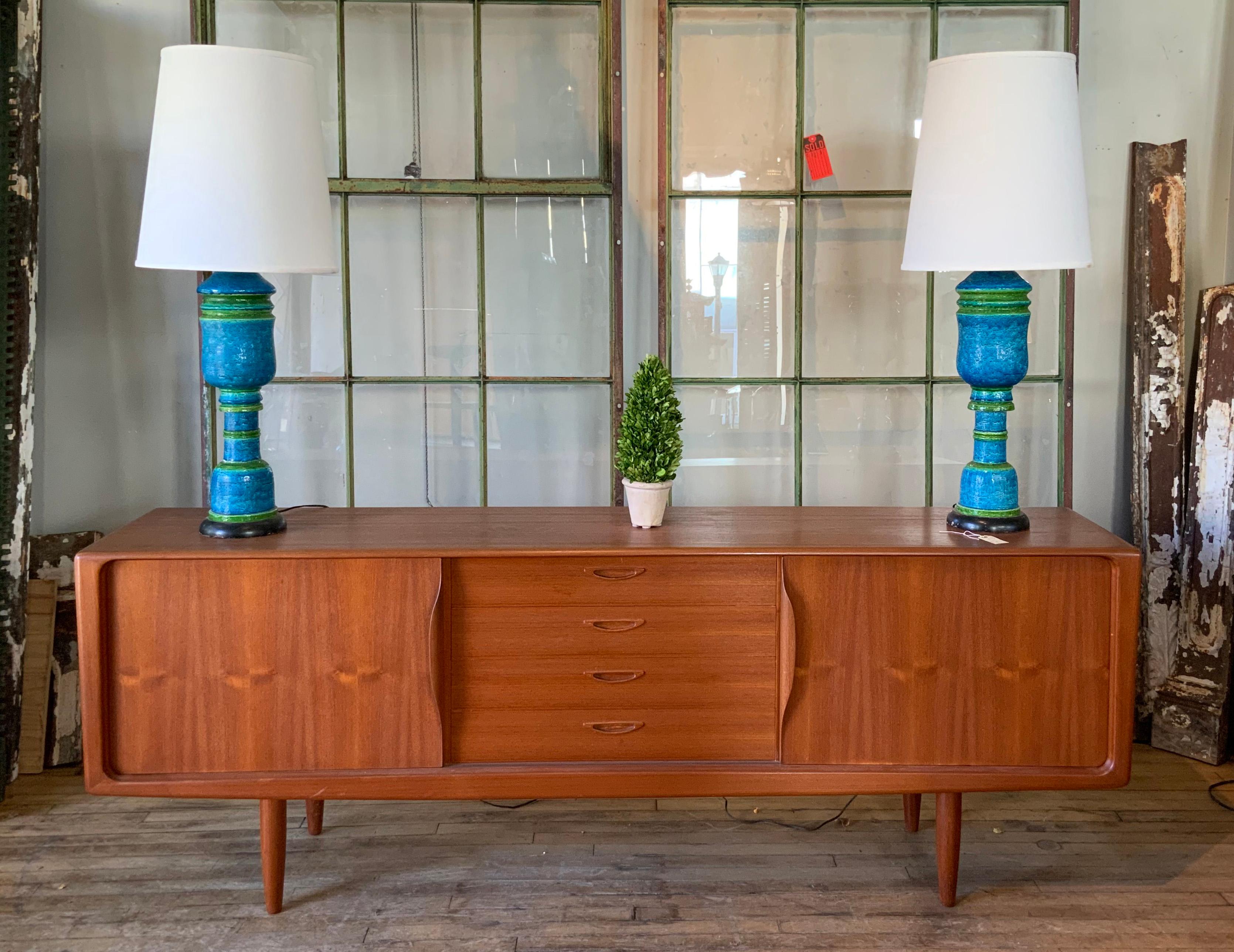 A beautiful and handsome 1950s Danish teak credenza cabinet, with sliding doors with integrated sculptural handles, which conceal adjustable shelves in the left and right side, and a bank of graduated drawers in the center. Raised on teak legs, this