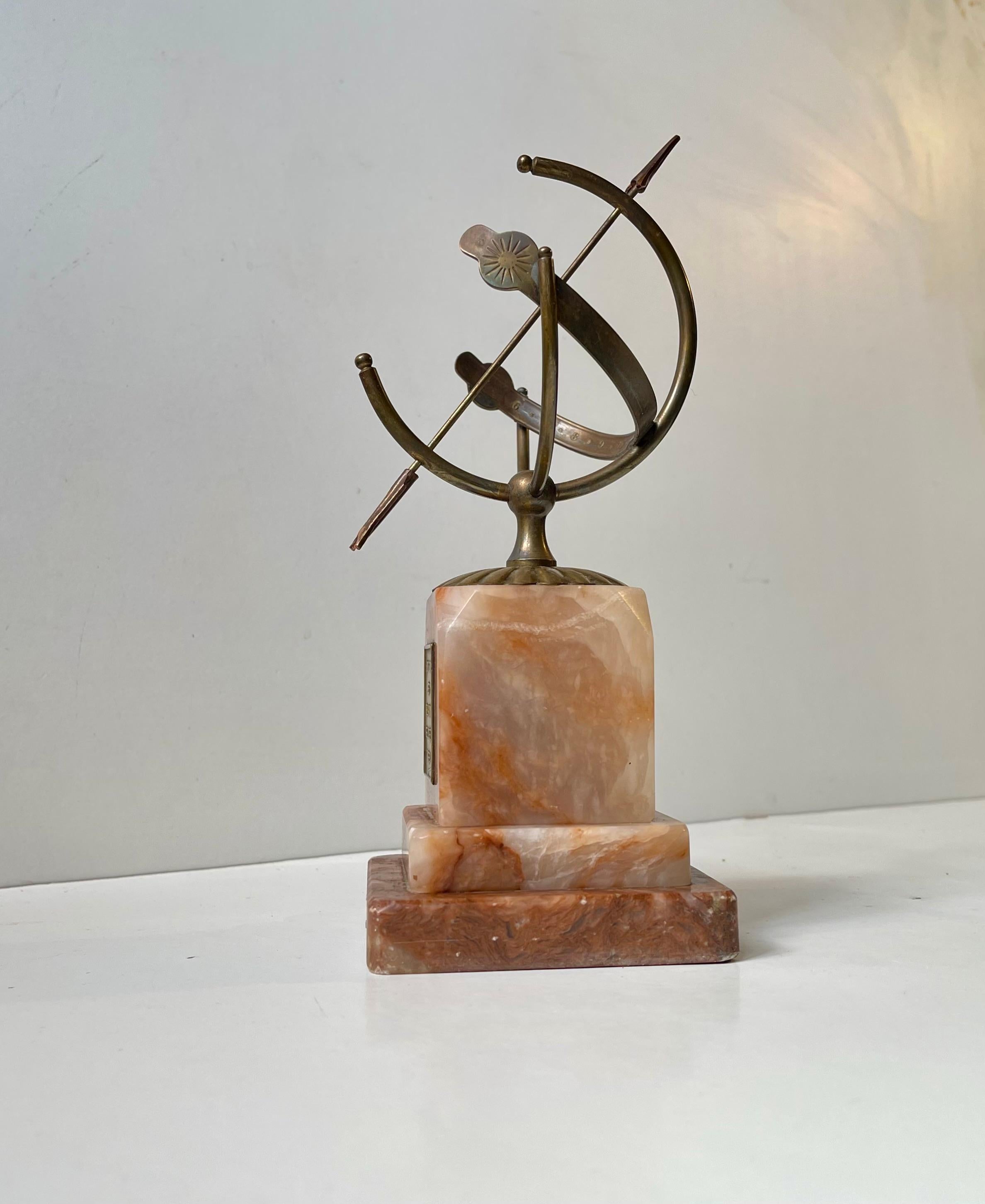Una elegante esfera armilar - reloj de sol elevada sobre dos bloques arquitectónicos de mármol ónice rojo/granate. Tiene una pequeña placa de bronce que dice en danés 