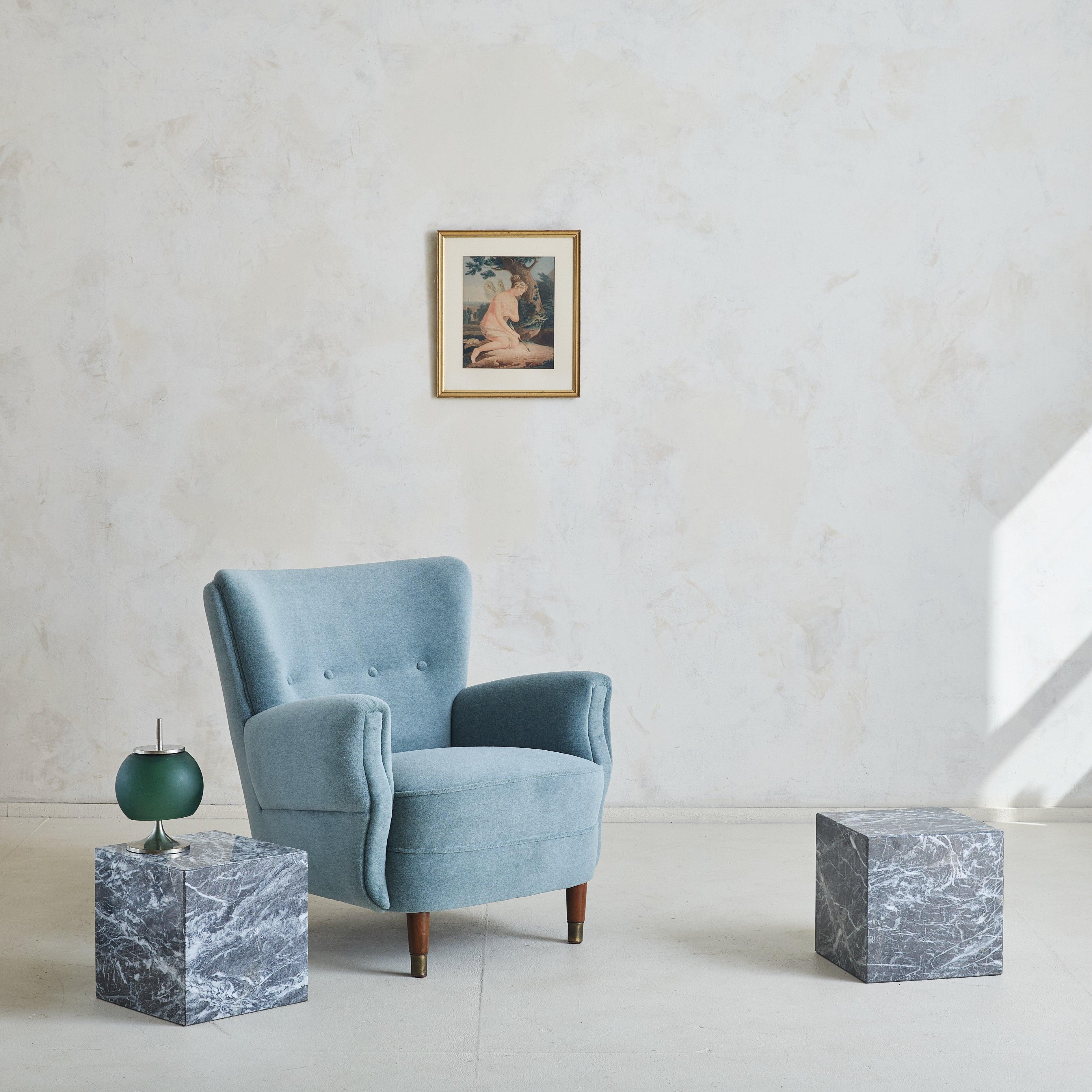 An inviting Danish armchair from the 1960s, carefully reupholstered in a soothing blue velvet. Its angular armrests and soft wingback create an effortless form, while tapered wooden legs capped in brass ground the piece. With its fluid lines and