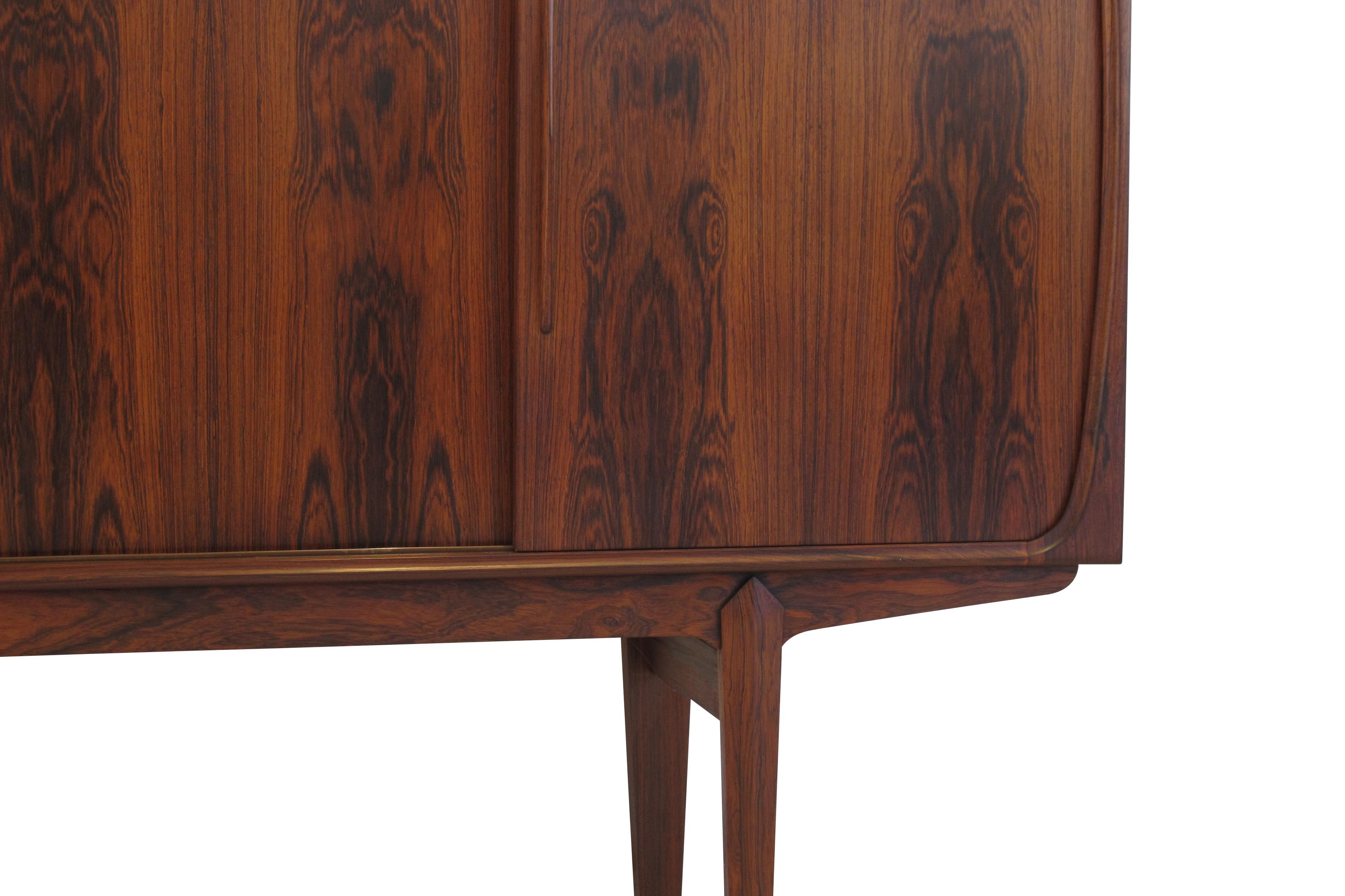 Brazilian rosewood credenza with figured book-matched grain raised on stilted legs. Four sliding doors with sculpted pulls reveal a mahogany interior with adjustable shelves. The middle section features a mirrored bar with eight silverware drawers