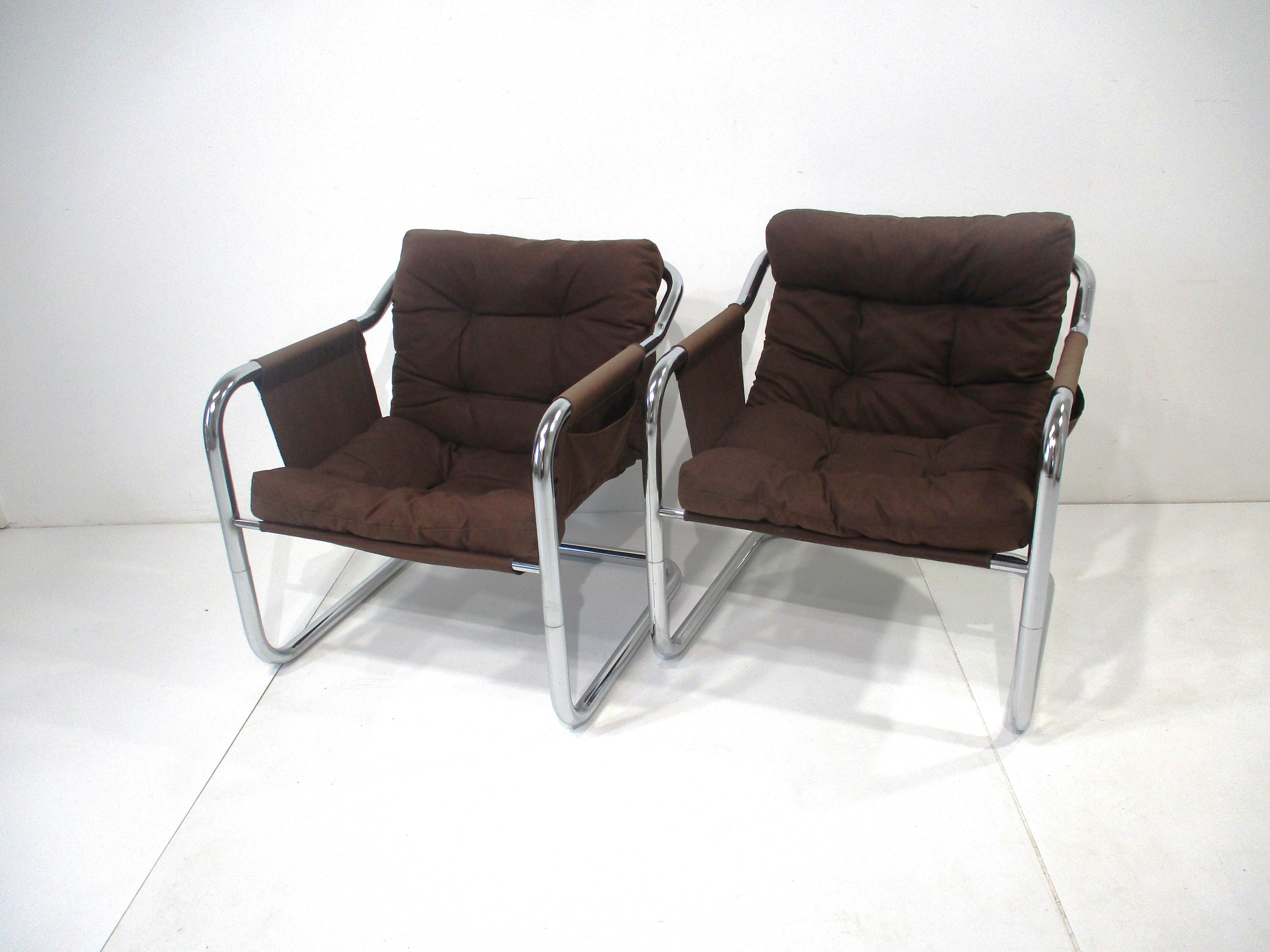 A pair of chromed steel sculptural tubed framed cantilevered lounge chairs with brown canvas styled sling and padded cushions . To each arm side there is a pouch for reading material or just for storage , retains the tag to one chair stating made in