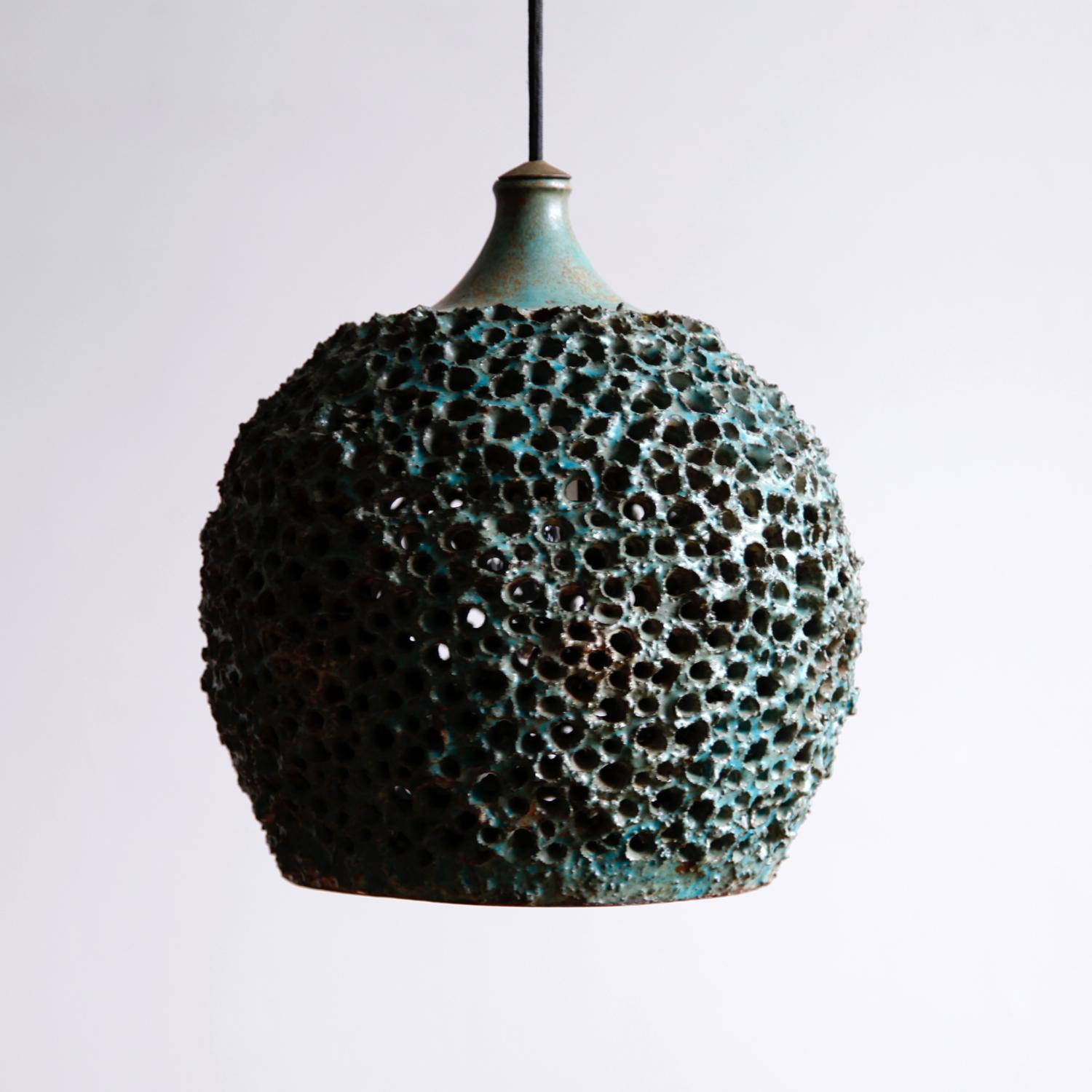 A beautifully made light fixture, resembling a sea urchin or other oceanic creature. The deeply textured surface is glazed with a color close to oxidized copper. Multiple perforations allow light to glow through the teal shade.