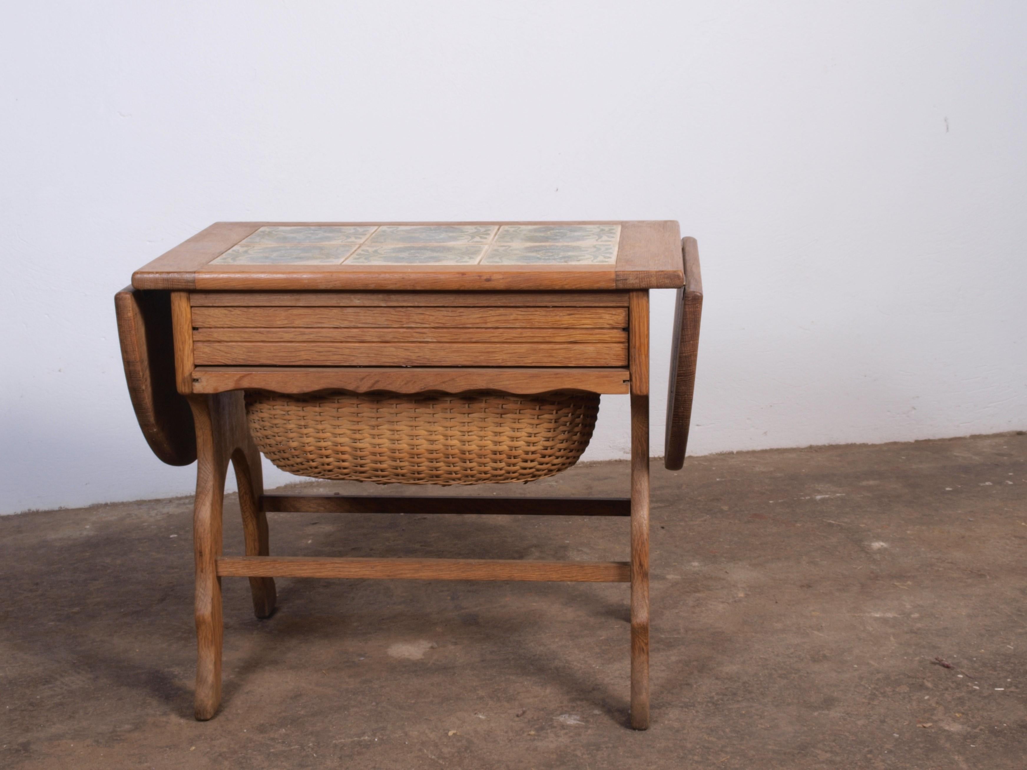 This Danish sewing table, crafted in the 1960s from sturdy oak with a tiled top, showcases solid craftsmanship. Featuring a drawer with storage and an extendable wicker basket, it adapts to various uses. The table's size adjusts from 60 cm to 84 cm,