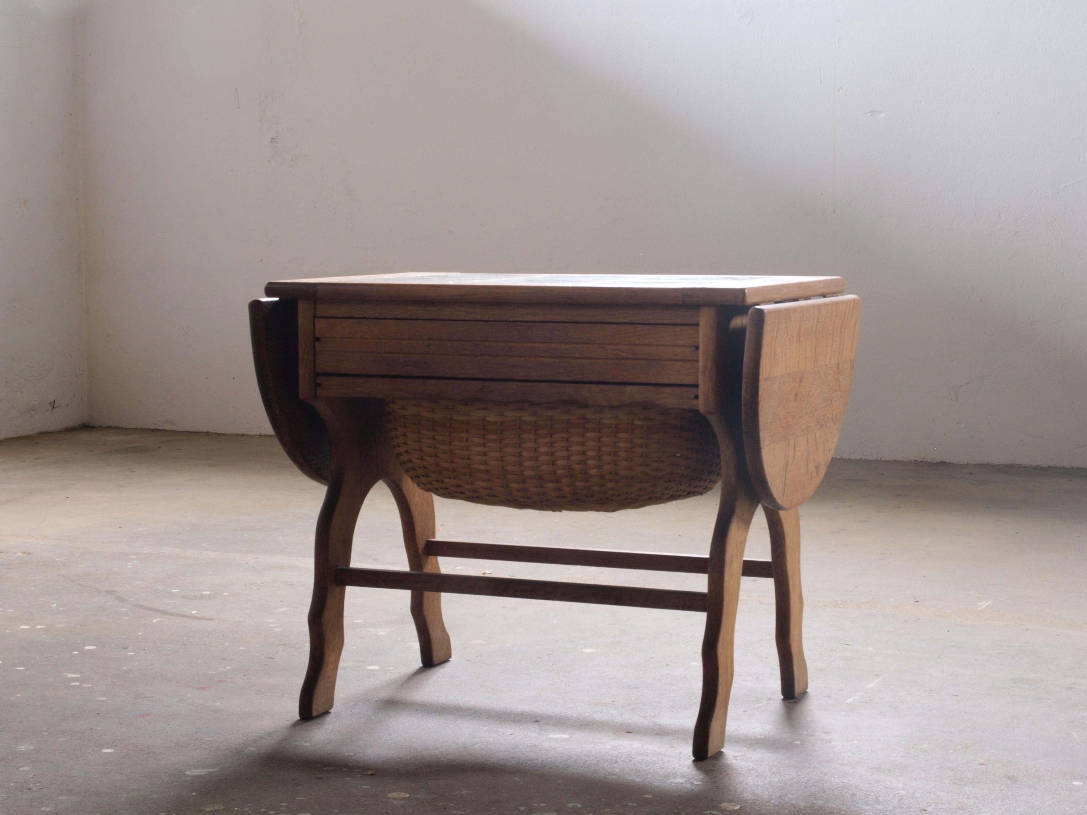 This Danish sewing table, crafted in the 1960s from sturdy oak with a tiled top, showcases solid craftsmanship. Featuring a drawer with storage and an extendable wicker basket, it adapts to various uses. The table's size adjusts from 60 cm to 84 cm,