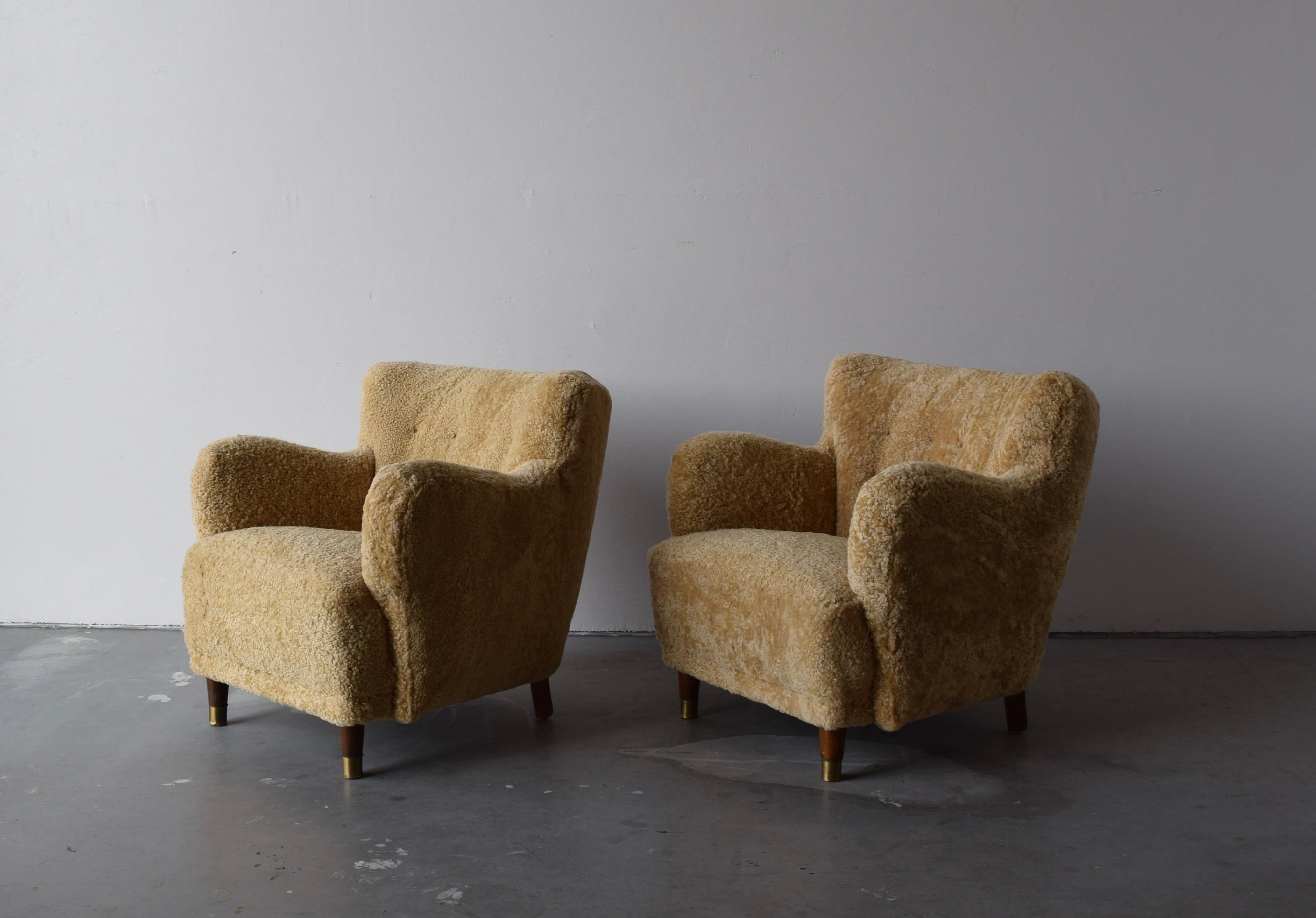 A pair of organic modernist lounge chairs. Designed and produced in Denmark, 1940s. Reupholstered in brand new authentic shearling upholstery. Dark stained wood, and brass caps to front legs. 

Similar in style to works by designers such as