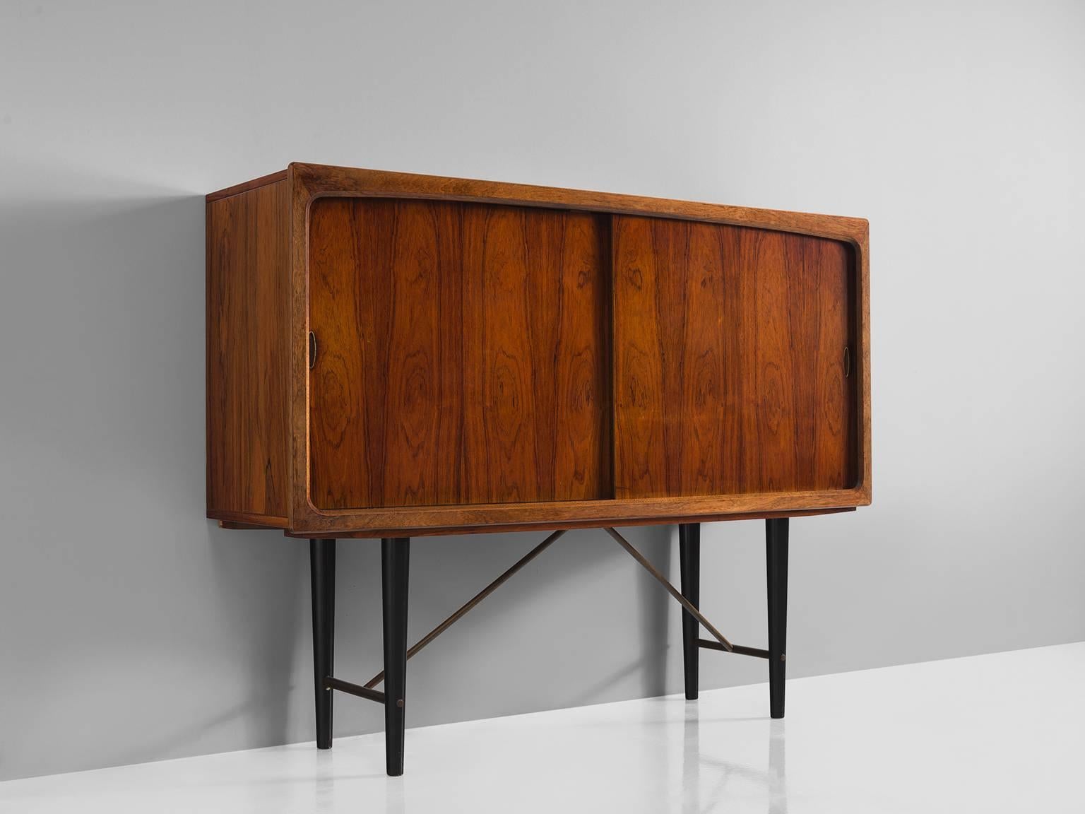 Cabinet, rosewood, black metal, Denmark, 1950s.

This cabinet is executed in rosewood and is typical for midcentury Scandinavian design. The cabinet features base with four black wooden tapered legs that are connected via horizontal rods and brass