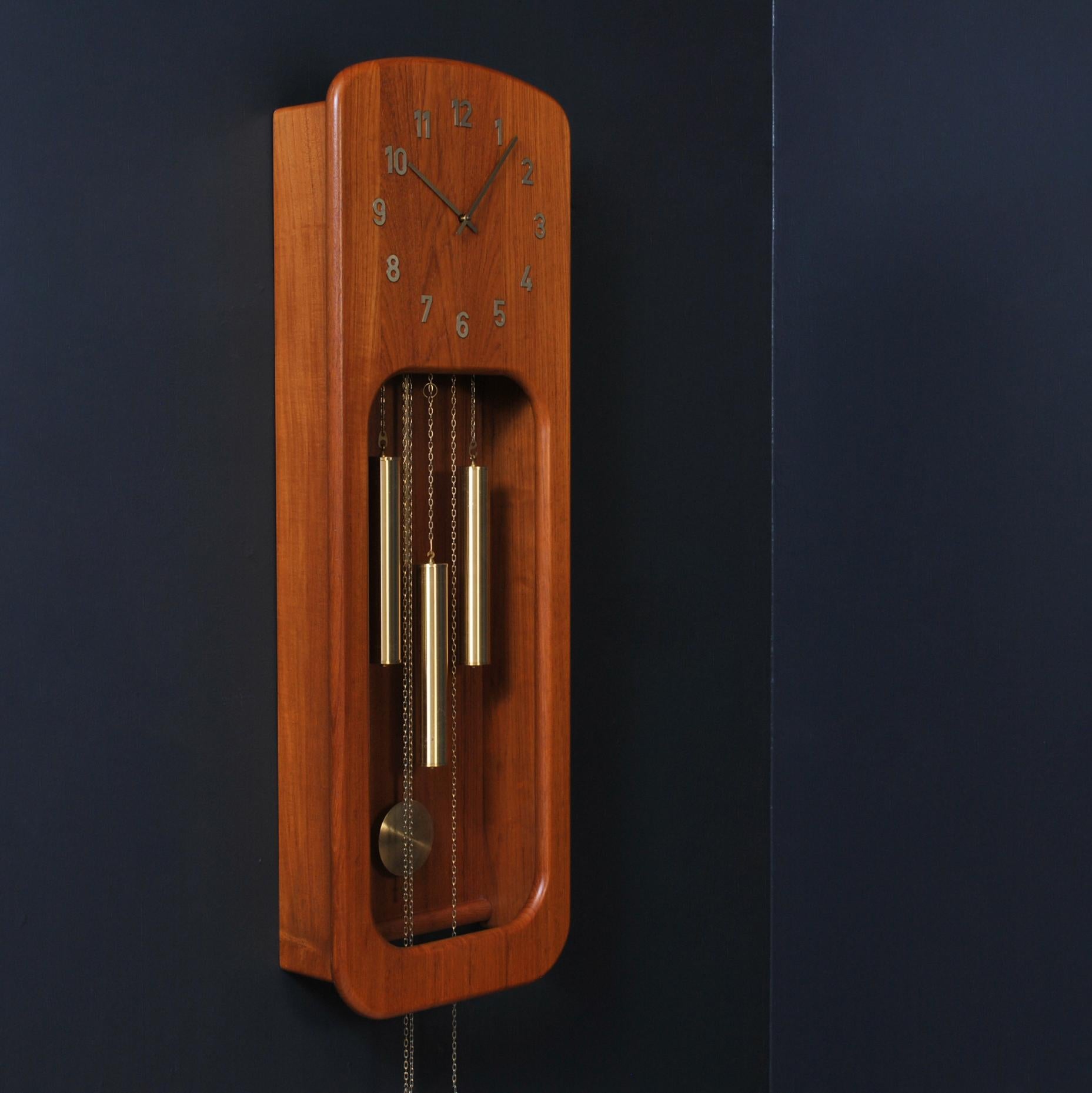 A large Danish long case chiming wall clock. Produced in Denmark circa 1960. A solid and bold design in teak with brass elements. A statement decorative Scandinavian modernist piece. Fully working and keeps perfect time. Time adjustable. The chimes