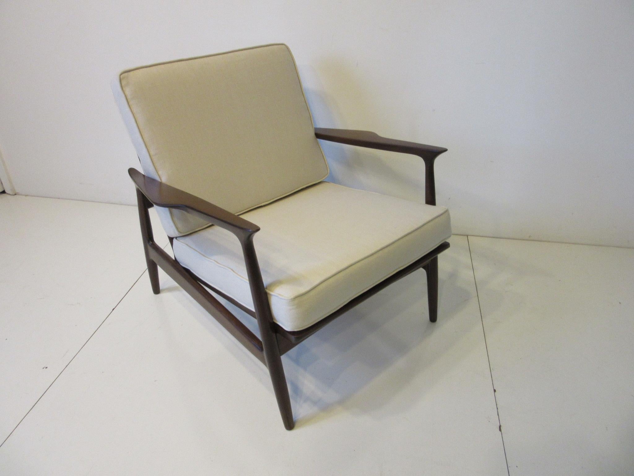A walnut finished wood framed lounge chair with sculptural arms and two upholstered cushions in a cream colored tight linen styled fabric. This well crafted piece has a painted raffia chair back with brass hardware giving it added detail, made in