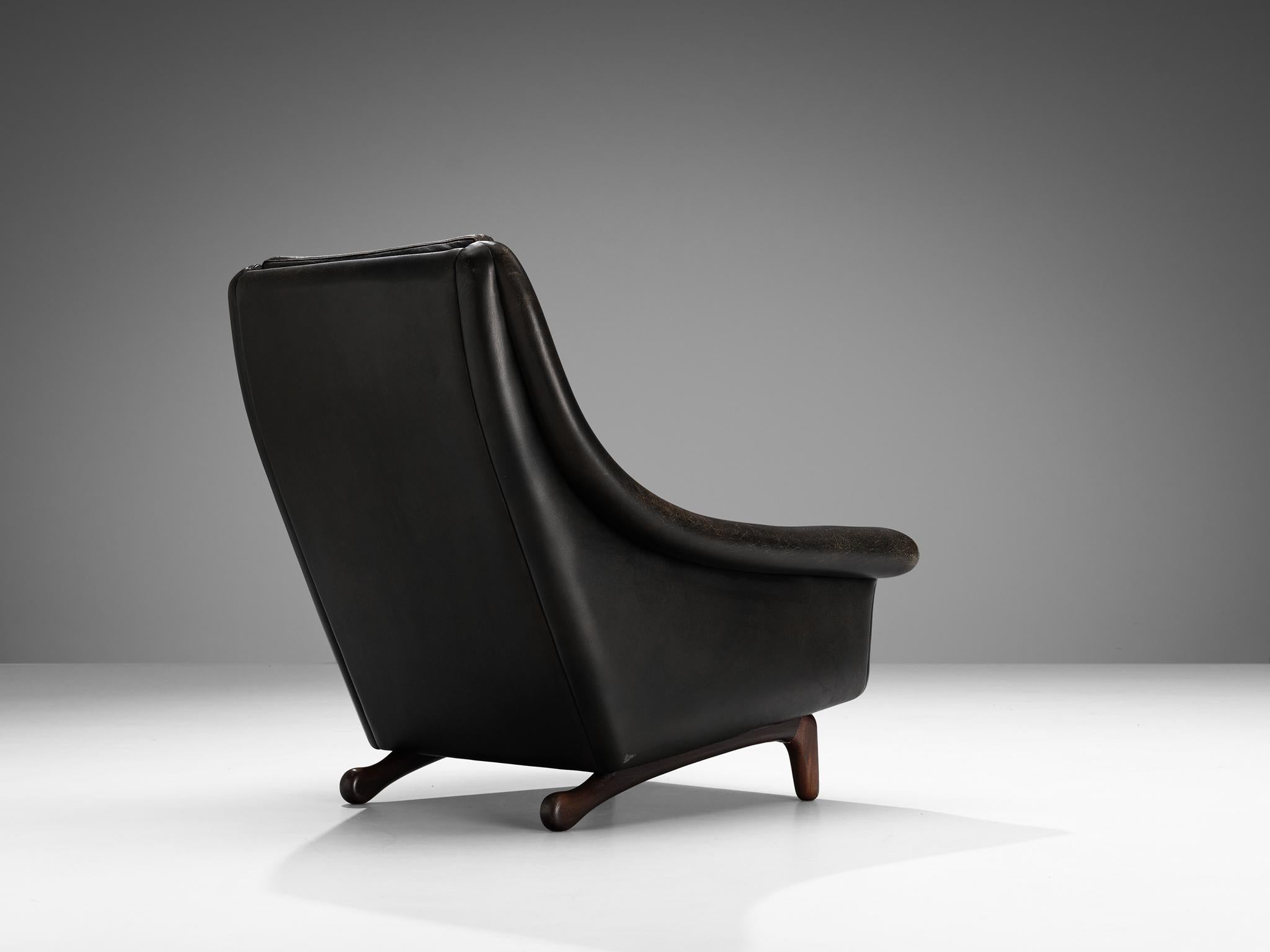 Fauteuil, cuir noir, teck Danemark, années 1960

Ce fauteuil aux formes organiques est doté d'une base modeste qui est sculpturale et fabriquée de manière exquise. Les pieds forment une excellente combinaison avec la forme courbée et fluide de la