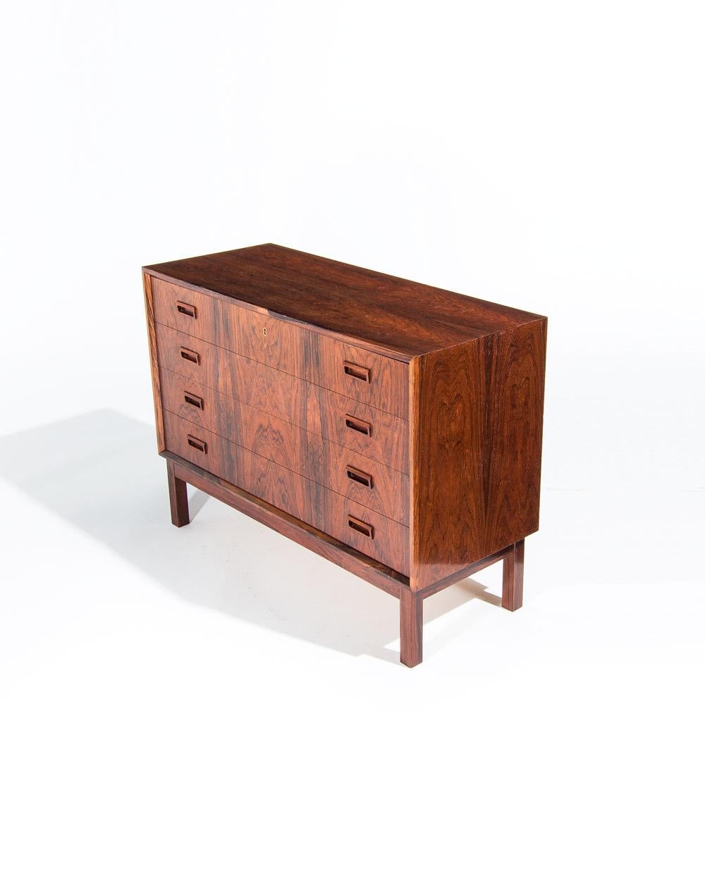 Esta cómoda danesa de palisandro de mediados de siglo, de la década de 1950, combina estilo y funcionalidad en un diseño compacto. Con cuatro cajones, el superior incluye cerradura y prácticos separadores para una mayor organización. Cuidadosamente