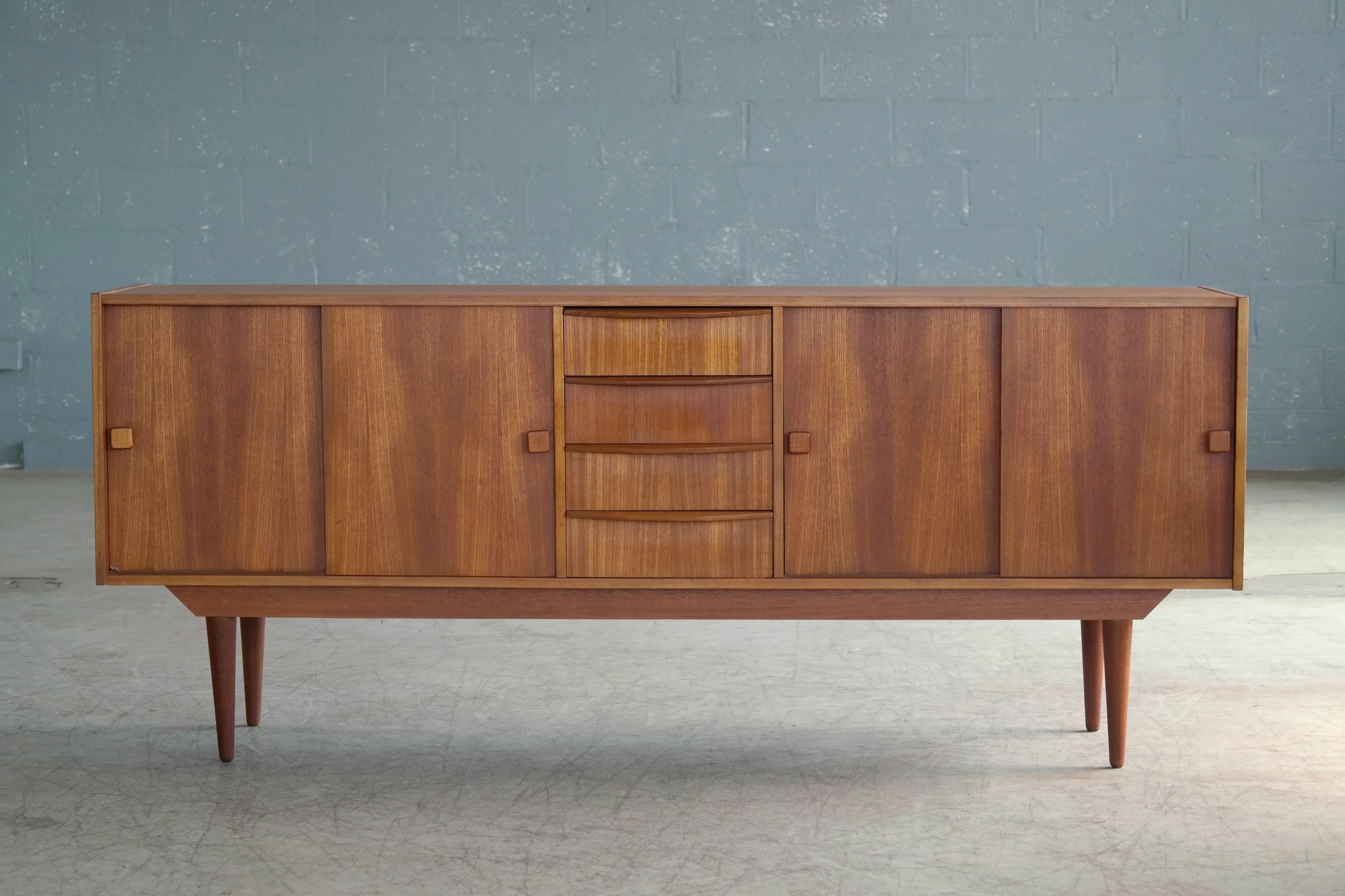 Beautiful Danish midcentury low sideboard in teak made by Domino Møbler in the 1960s. Nice center drawer section flanked by compartments with adjustable shelves behind nice bookmatched sliding door with carved pulls. Very nice color and grain and