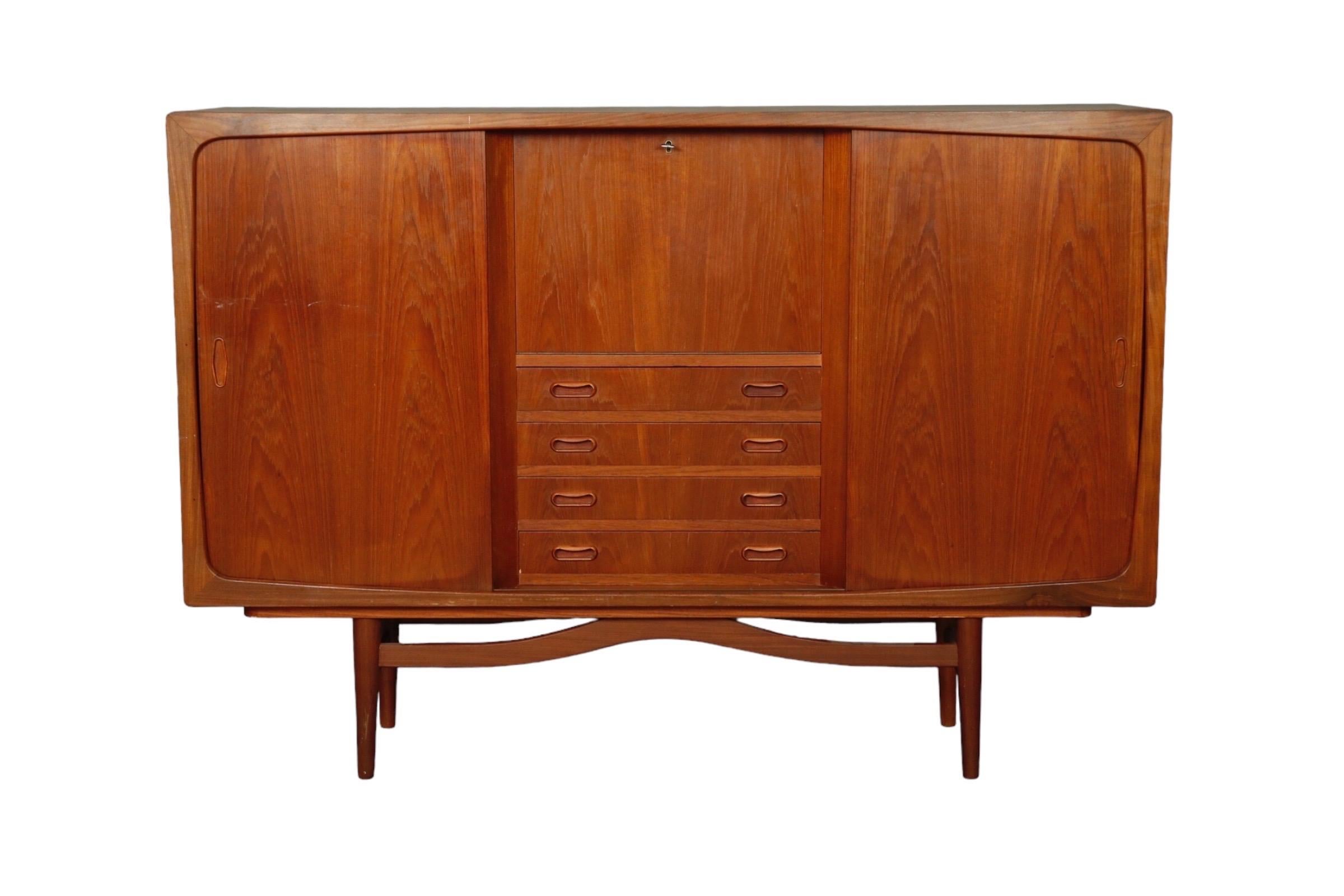 A Danish mid century modern buffet in teak. A central drop front reveals a mirrored bar area with a glass shelf. Below are four drawers with kidney shaped recessed handles. Sliding doors on each side decorated with bookmatched veneers house storage