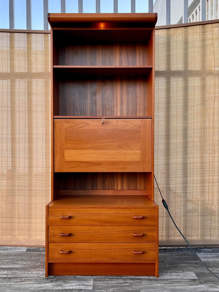 Vintage Danish Mid Century Modern Drop Down Secretary Desk Cabinet by Domino Mobler. Circa 1970s.
Features a beautiful teak wood grain, a work surface via the secretary desk compartment, as well as plenty of additional storage with three drawers at