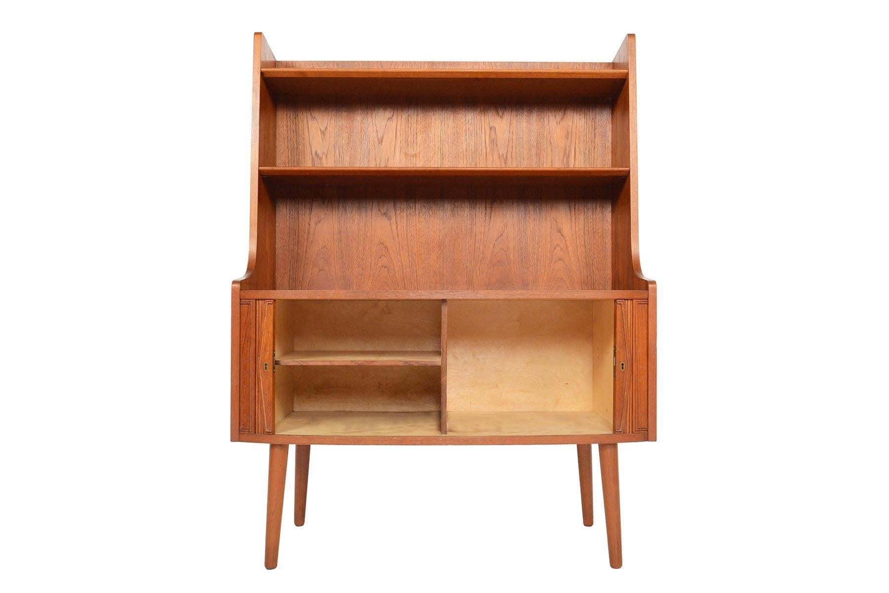 This Danish modern teak bookcase offers an unusually short case with a beautiful bow front design! The bookcase back offers an upper fixed shelf and a middle adjustable shelf. The tambour doors scroll open to display two bays one of which is