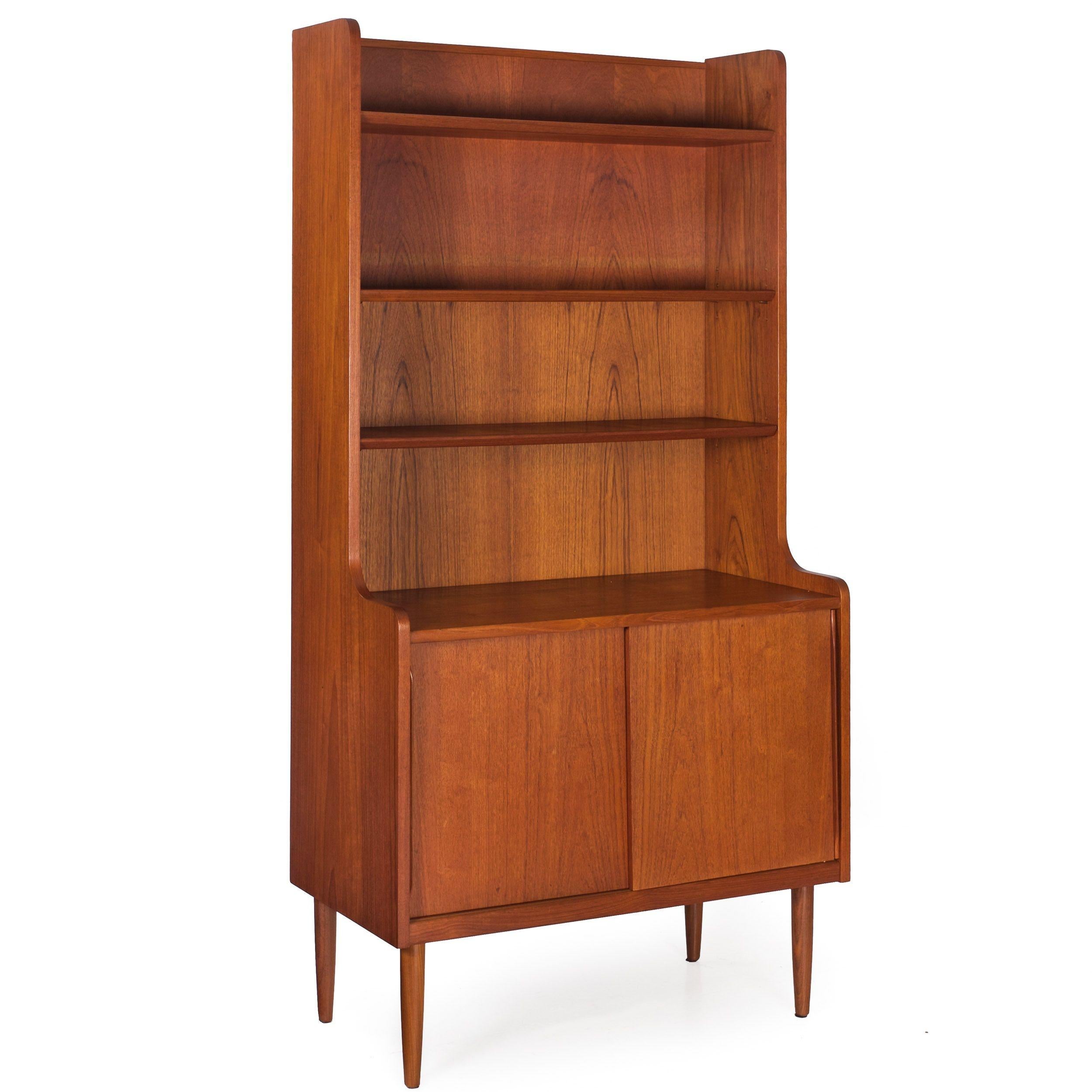 A sleek Danish Modern teak cabinet with three adjustable open shelves over an enclosed cabinet with sliding doors and molded handles. The cabinet is crafted in one integral piece and rests over turned and tapered legs. An old partial label is