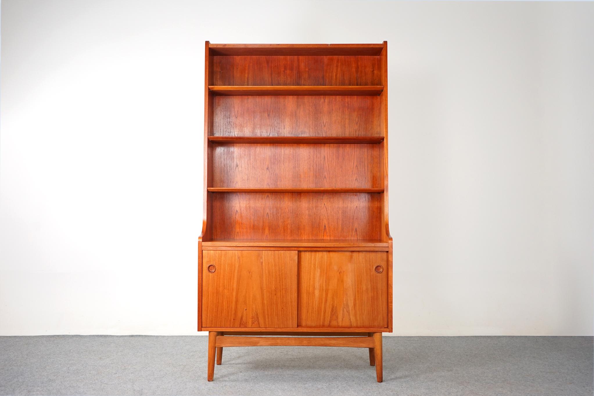 Teak Danish bookcase, circa 1960's. Tapered open bookcase with three adjustable shelves, graduating in depth. Lower cabinet with two sliding doors and 1 adjustable shelf within. Case sits upon removable solid wood base with tapering legs and a