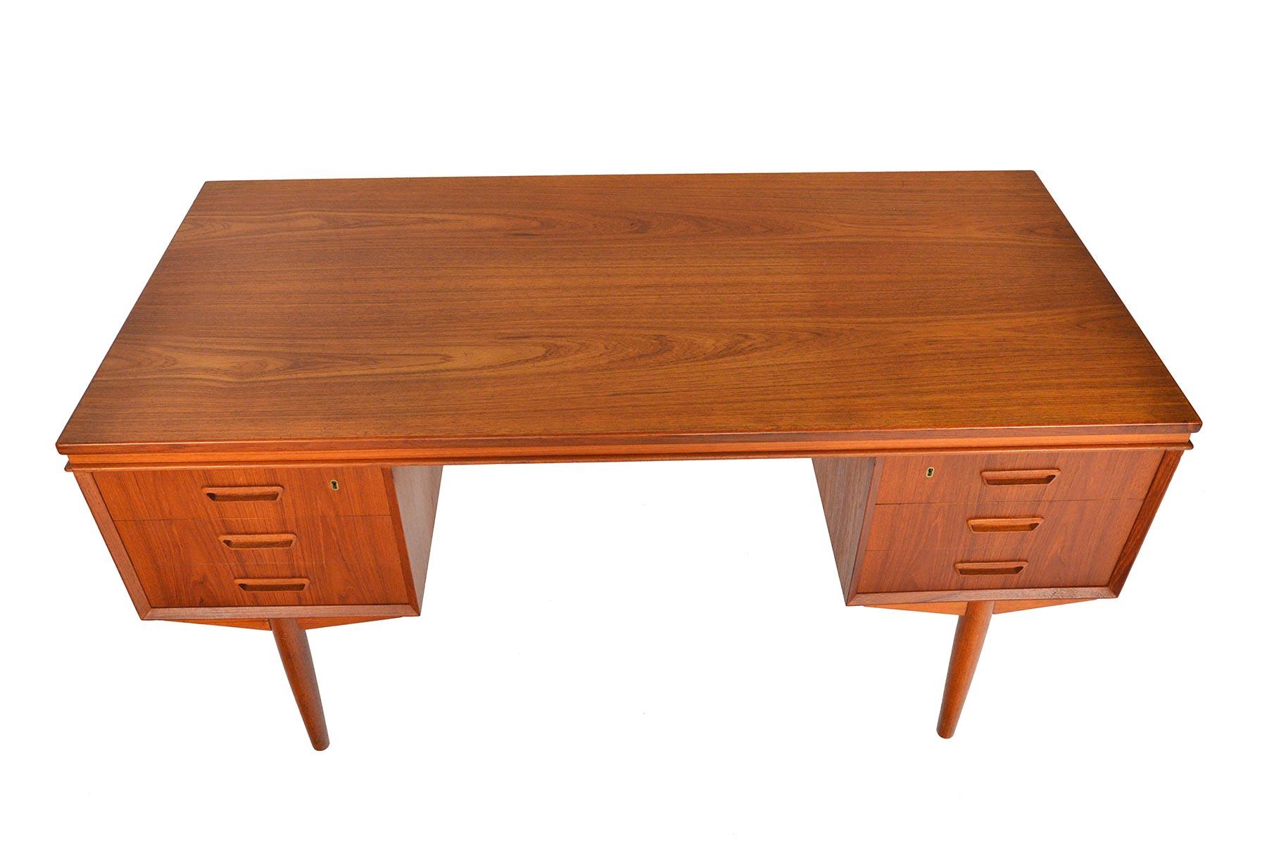This Danish modern teak executive desk provides a wide work surface supported by two banks of drawers standing on spindle legs. Each drawer is adorned with a carved trapezoid pull. The back features divided bookshelf storage. This desk offers a tall