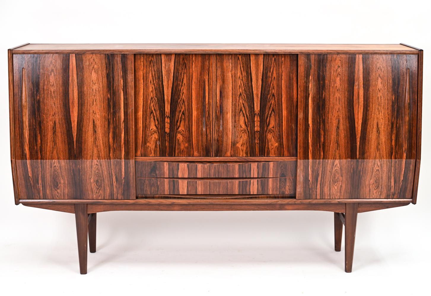 A stunning sculptural Danish mid-century tall bar sideboard or credenza in handsome rosewood veneer, featuring a geometric angled front on tapering legs. Sliding cabinet doors that open to reveal ample shelving flank a central bar cabinet with an