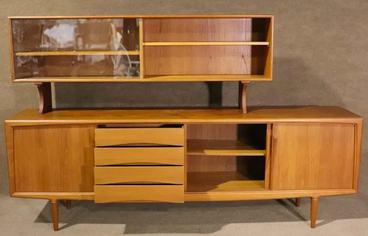 Danish modern design in the style of Arne Vodder, featuring a long teak credenza with cabinet and drawer storage. Topper with glass doors, sculpted door handles, tapered legs.
Please confirm location.
