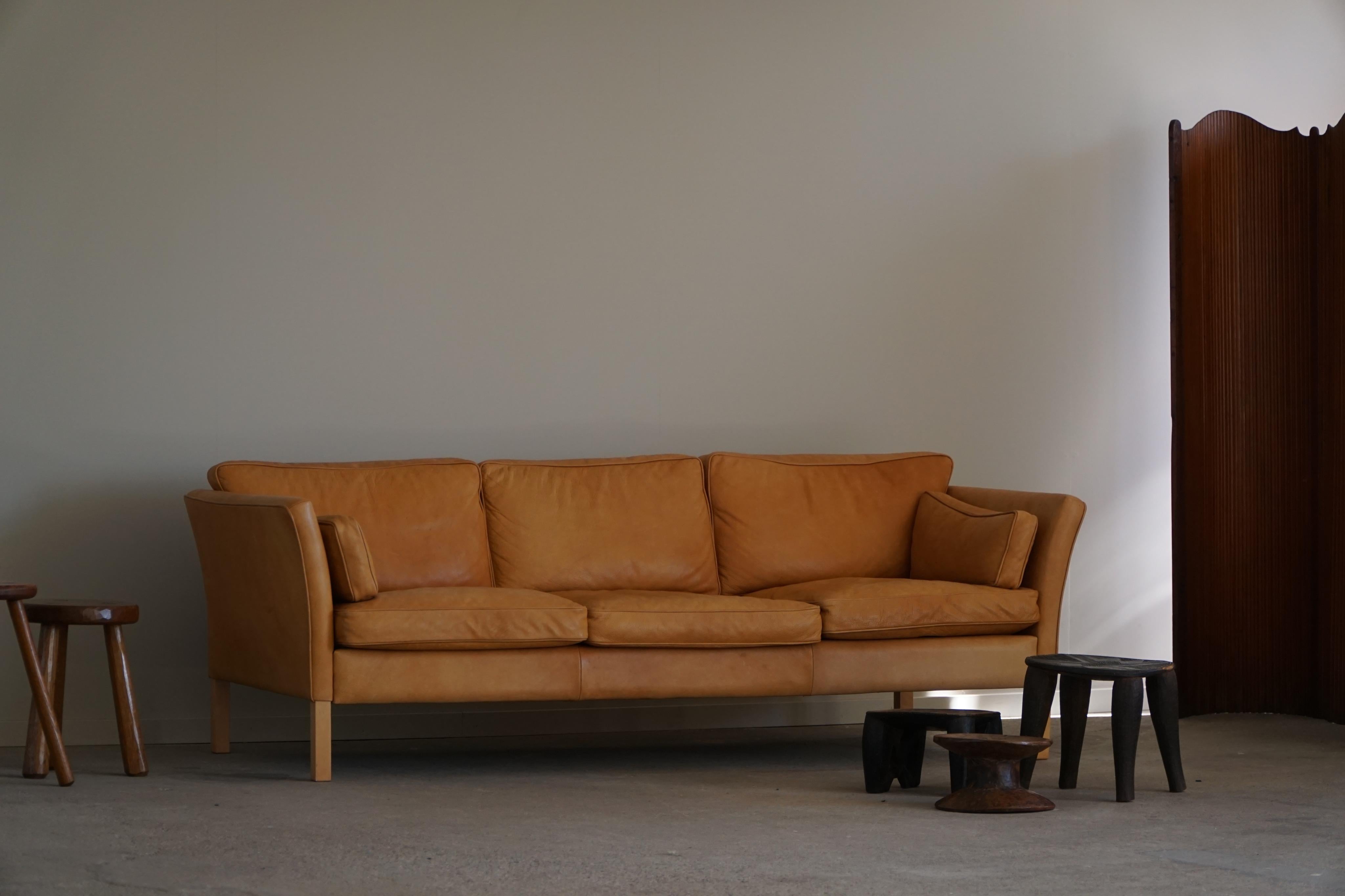 Un très beau canapé trois places classique de style moderne danois, fabriqué par Stouby au Danemark. Le canapé est revêtu d'un cuir brun cognac de haute qualité, dont la patine riche et chaude lui confère un aspect vintage et luxueux. Les pieds sont