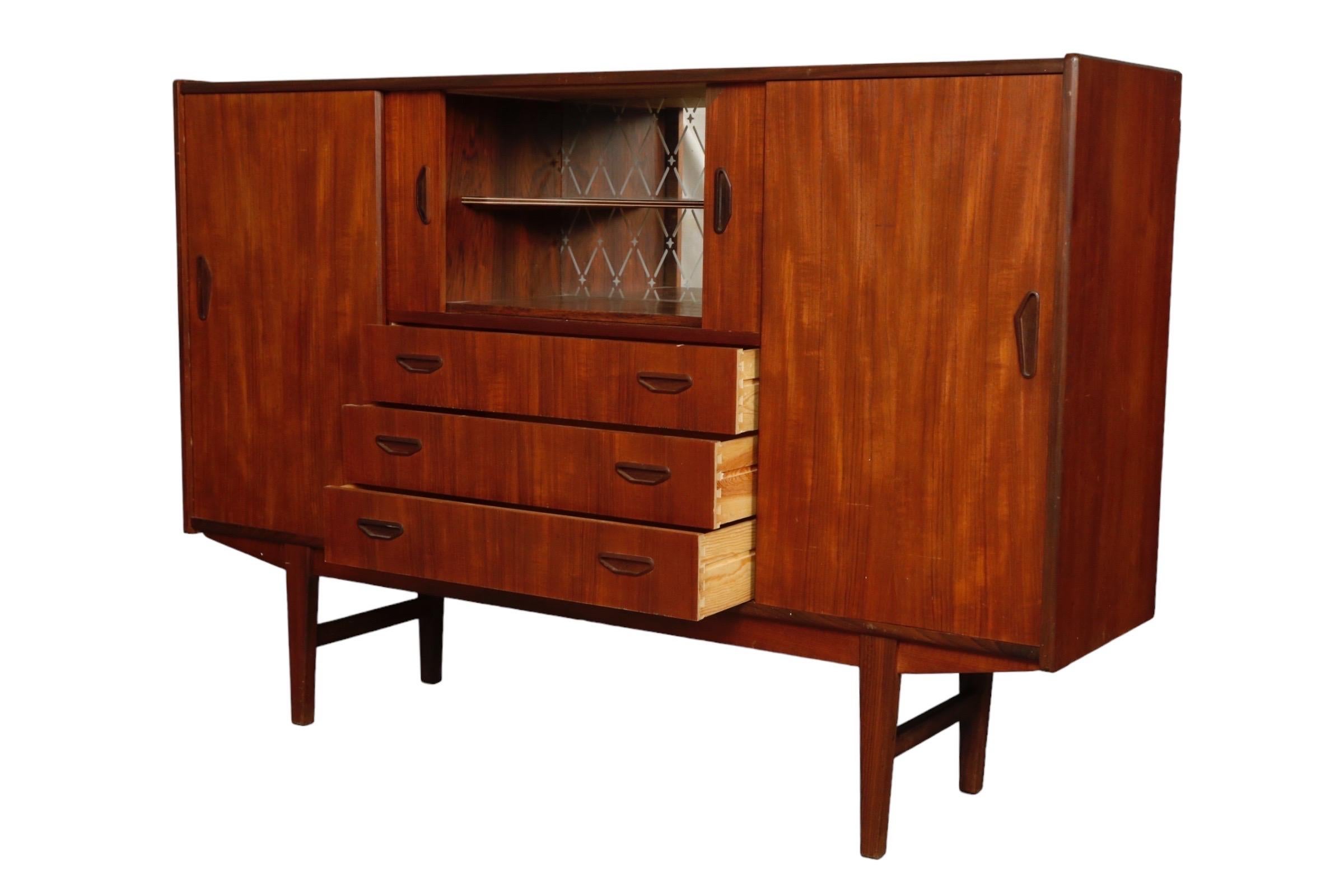 A Danish mid century tall buffet. Small sliding doors above three dovetailed drawers in the center open to reveal a mirrored bar area. On either side larger sliding doors reveal six shelves and ample storage. Finished with round tapered legs.