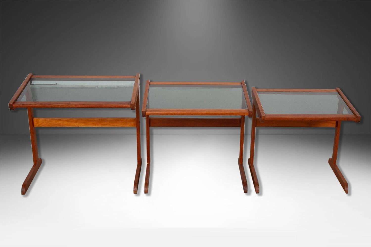 Set of three Mid Century Modern teak, rectangular smoked glass top nesting tables. The top rests on an attractive trestle base with a crossbar. Exceptionally well crafted.  Undersides of the tables are channeled so that the tables hook to one