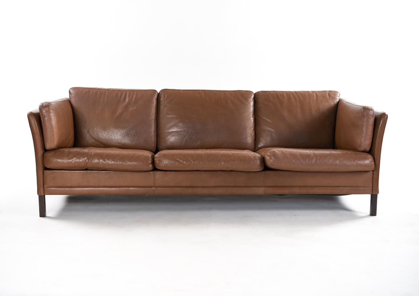 A beautiful Danish midcentury sofa in caramel leather which can seat three comfortably. Made by Mogens Hansen, this sofa is a wonderful example of modern design with subtle lines and form. The leather is showing an attractive patina from age.