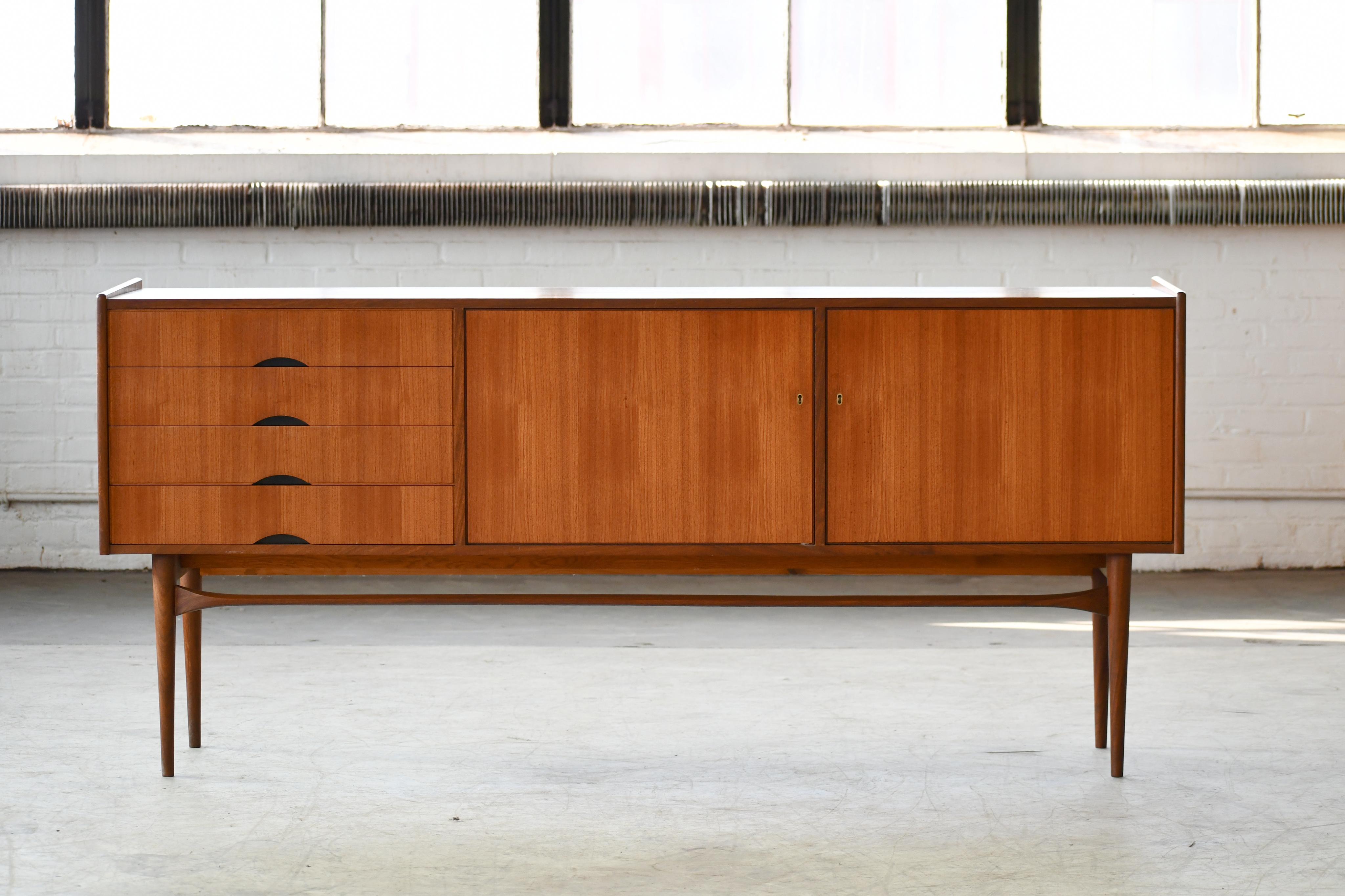 Beautiful Danish midcentury low sideboard in teak made in 1960s. Nice off-center drawer section with room for cutlery flanked by compartments with adjustable shelves behind nice book matched sliding doors with carved pulls. Raised on thin elegant