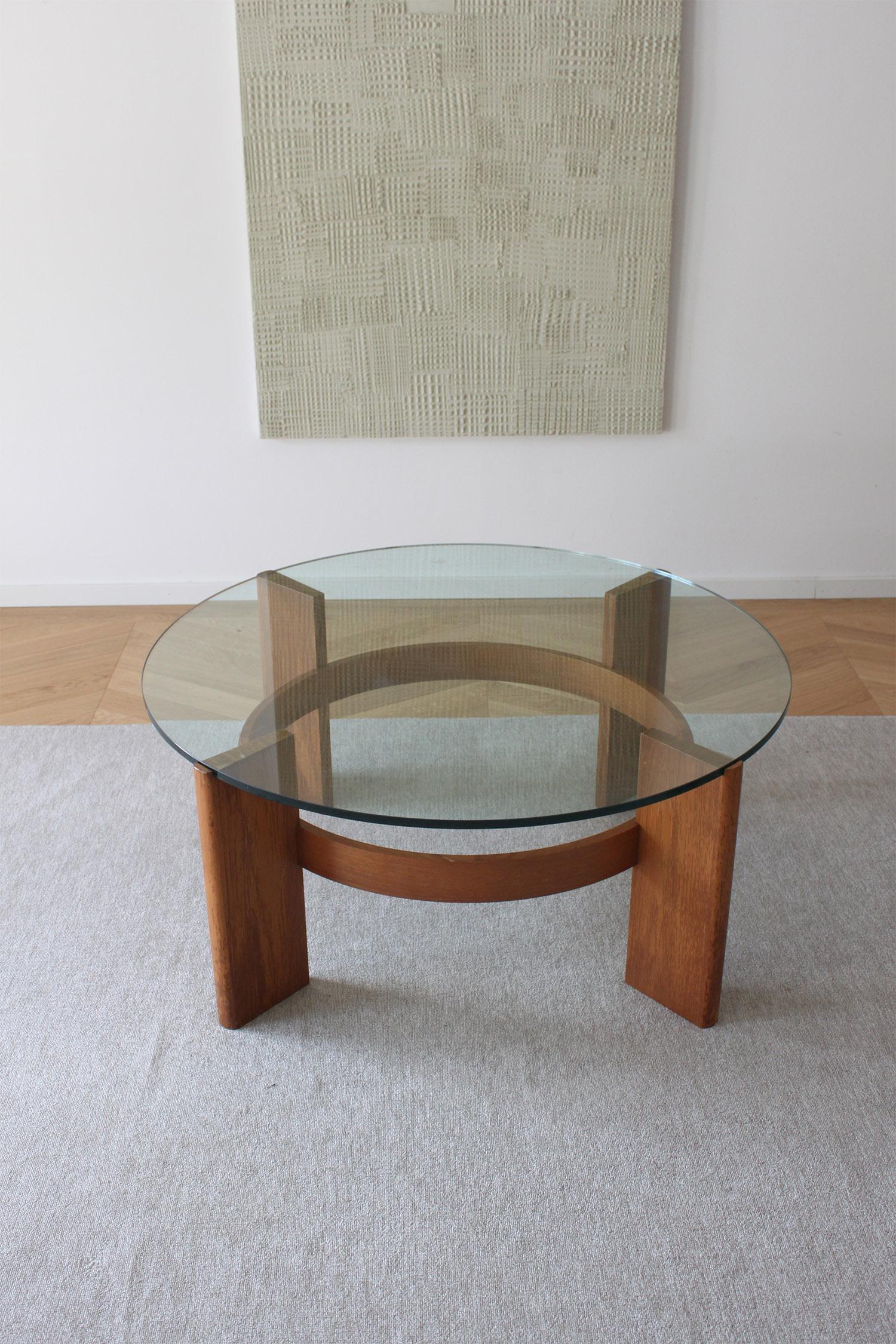 Esta es una clásica mesa de centro danesa moderna de mediados de siglo, con un bonito tablero circular de cristal y una robusta base de madera. 
El diseño de la mesa encarna la elegancia minimalista de los muebles escandinavos, con líneas limpias y