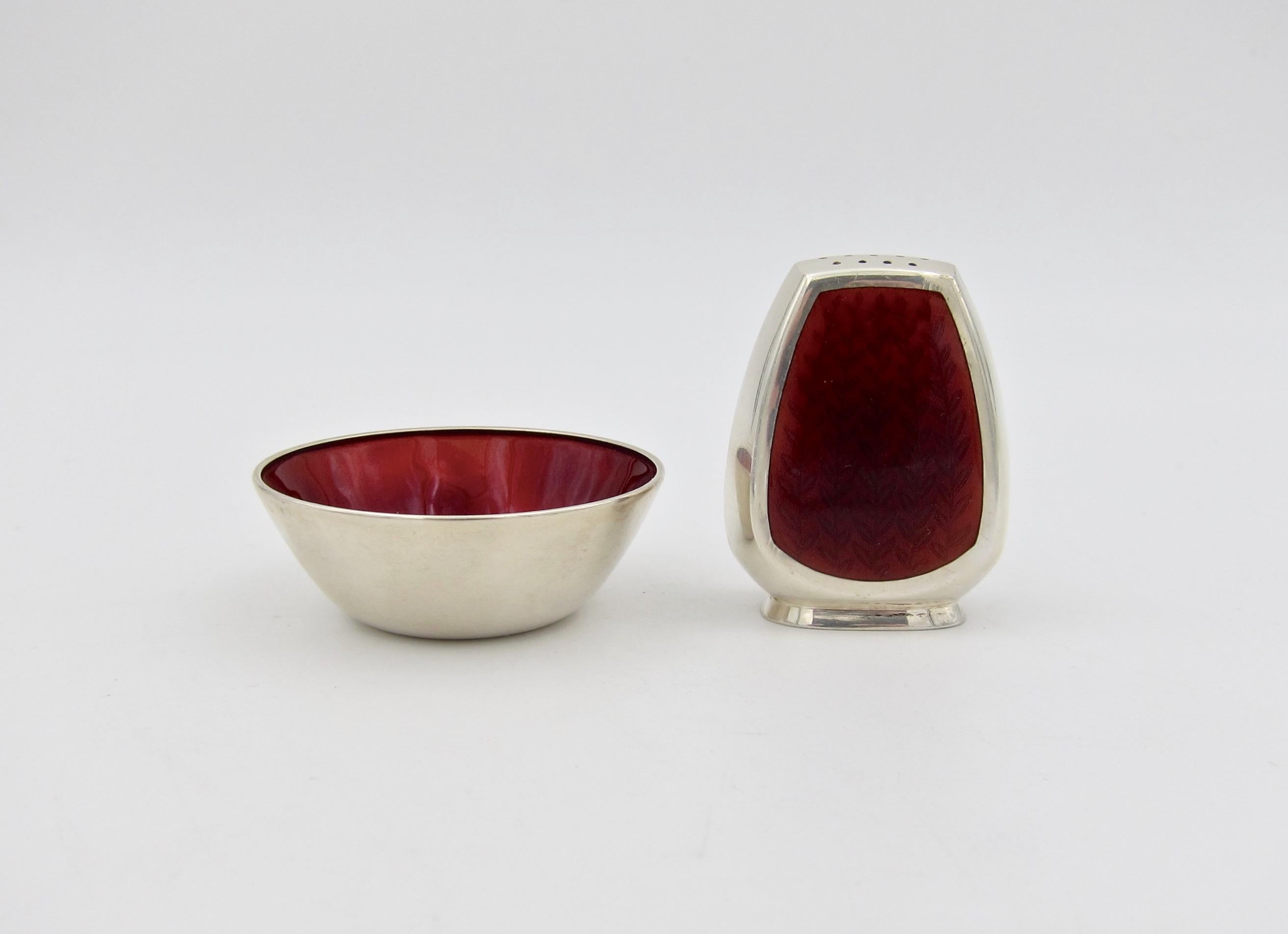 A Danish midcentury open salt cellar and pepper Shaker in sterling silver decorated with maroon red enamel from Anton Michelsen silversmiths of Copenhagen, dating circa 1950. The set consists of a salt dip with enamelwork over a floral motif while