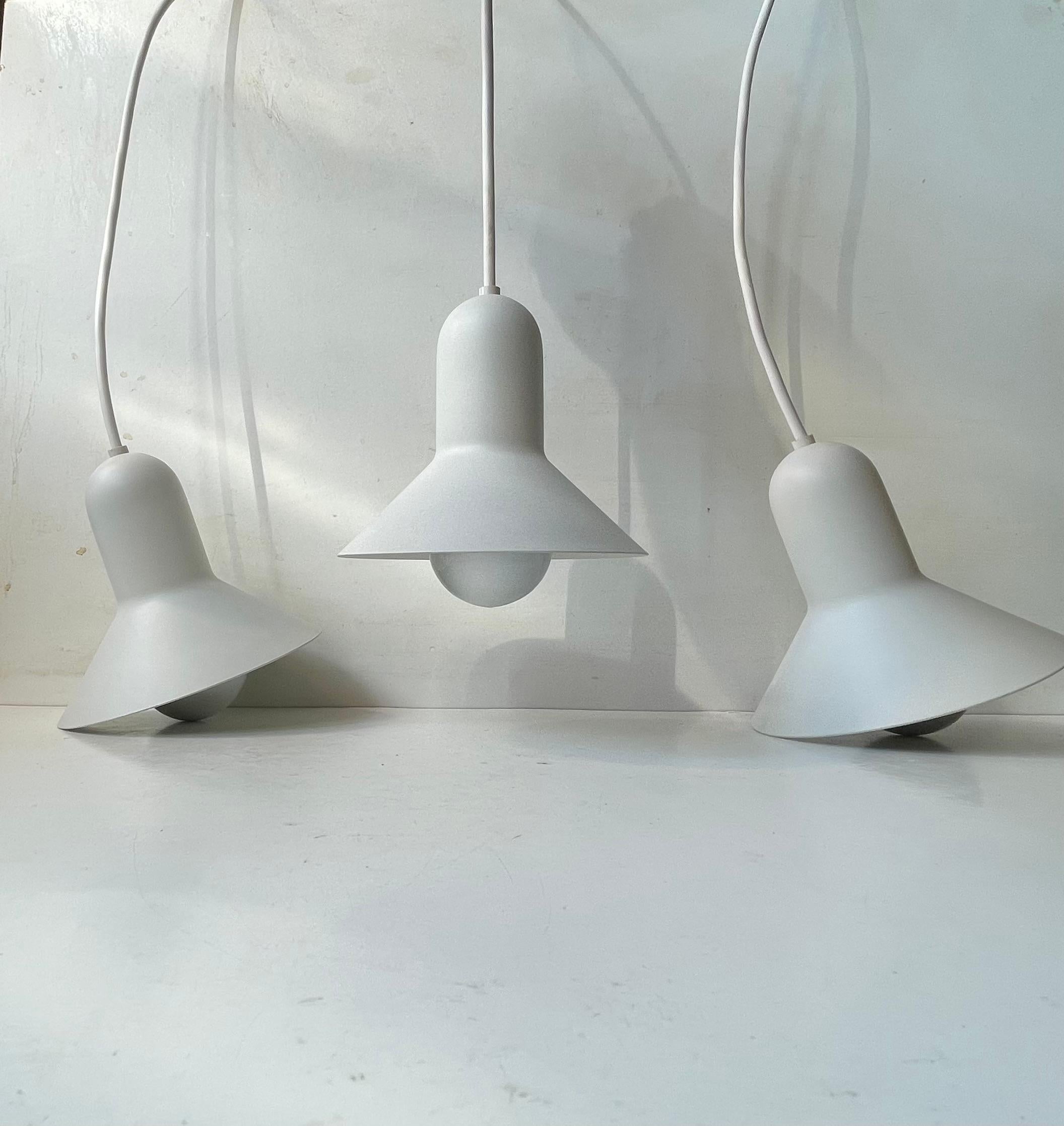 Un trío de pequeñas lámparas colgantes blancas de metal blanco. Estricta apariencia minimalista combinada con curvas orgánicas. Este modelo se llama Confetti y fue diseñado por el arquitecto danés Claus Bonderup y fabricado por Focus en Dinamarca a