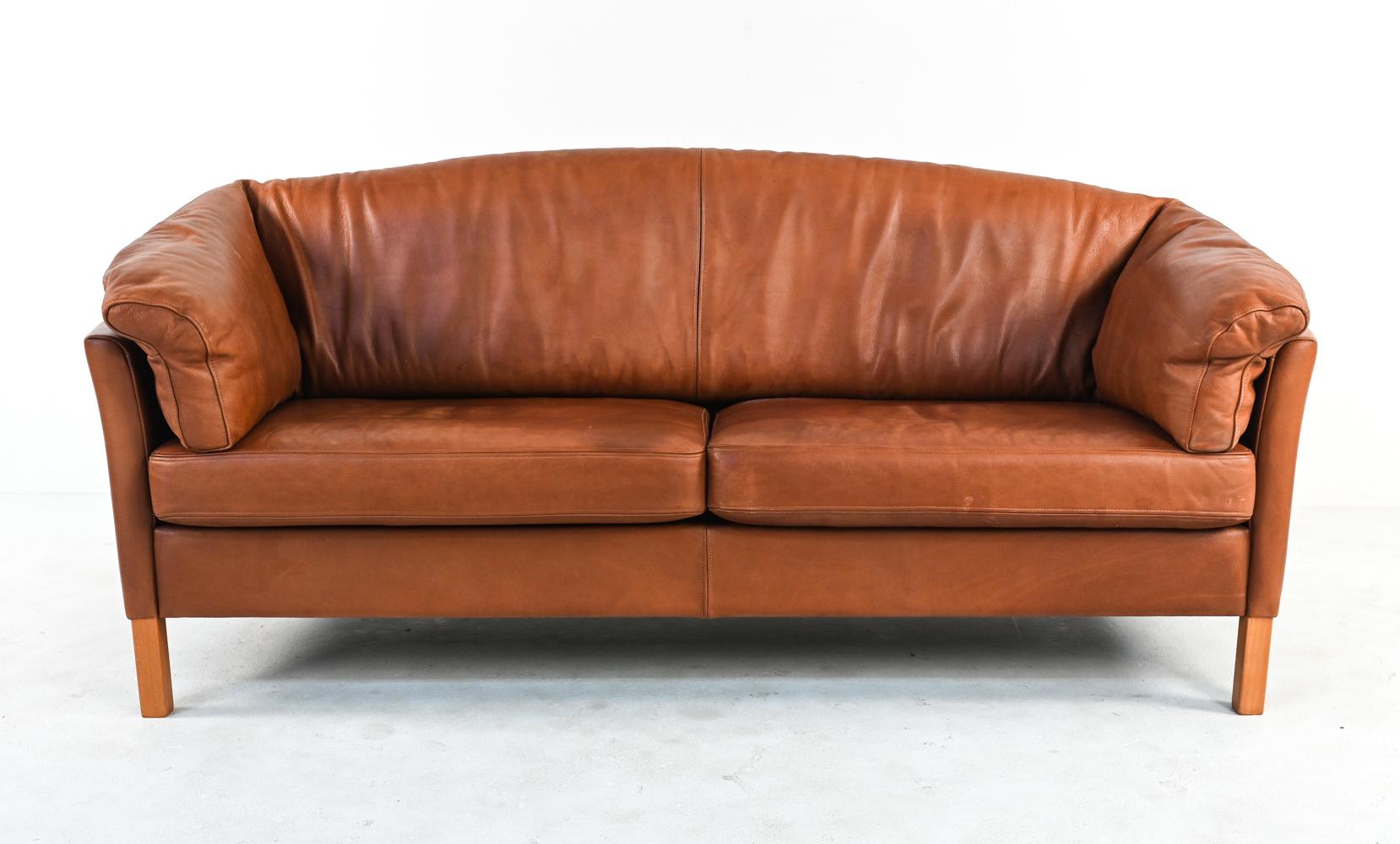 This iconic model 535 two-seat sofa by Mogens Hansen features slightly bowed shelter arms and comfortable plush cushions atop sturdy natural beech wood legs. The upholstery is a handsomely patinated tan leather - reminiscent of an English saddle -