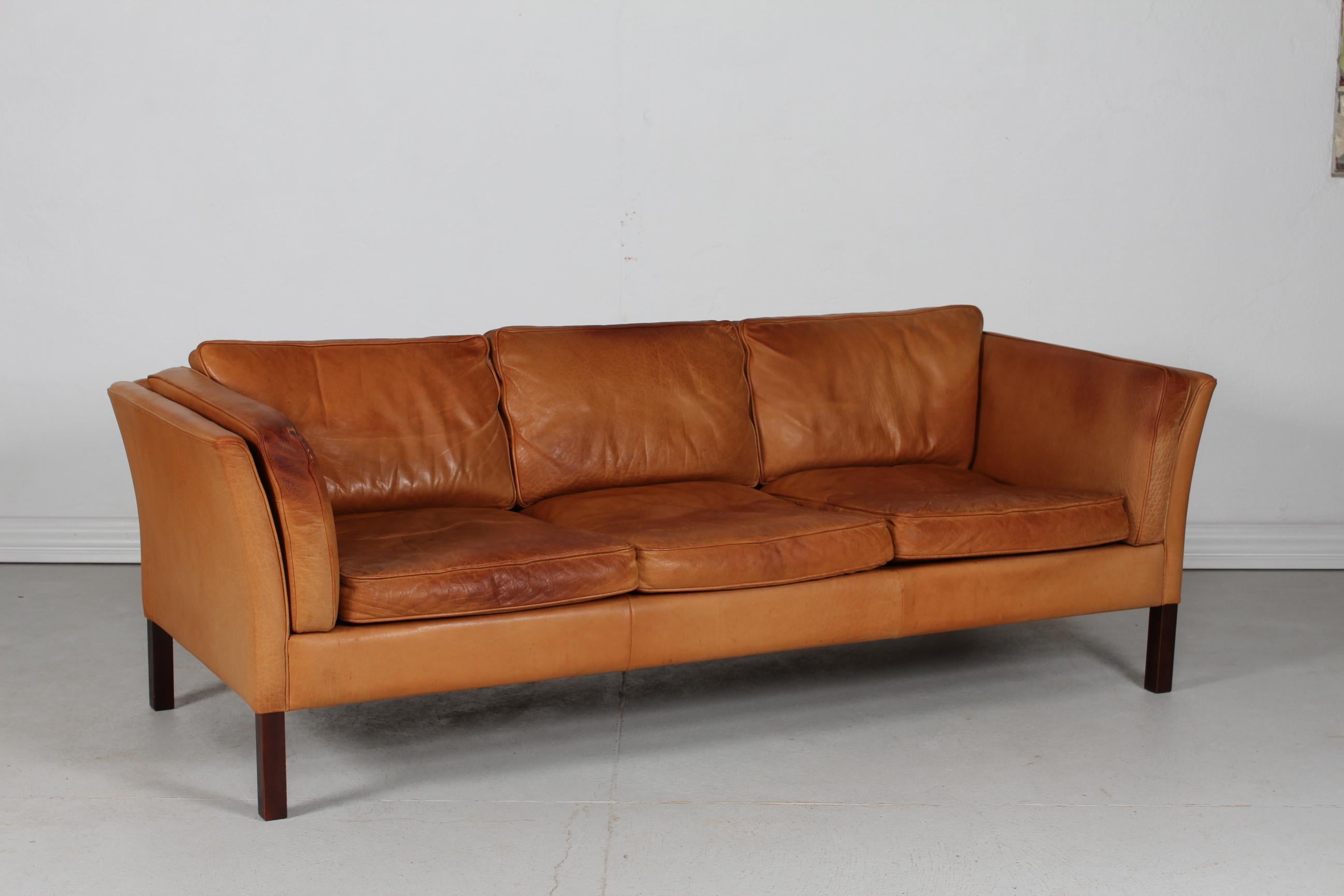 Danish vintage 3-seater sofa in the style of Børge Mogensen.
This sofa is manufactured by Stouby Møbelfabrik in Denmark in the 1970s
It's upholstered with the original cognac-colored leather with good patina. 
The square legs are made of dark