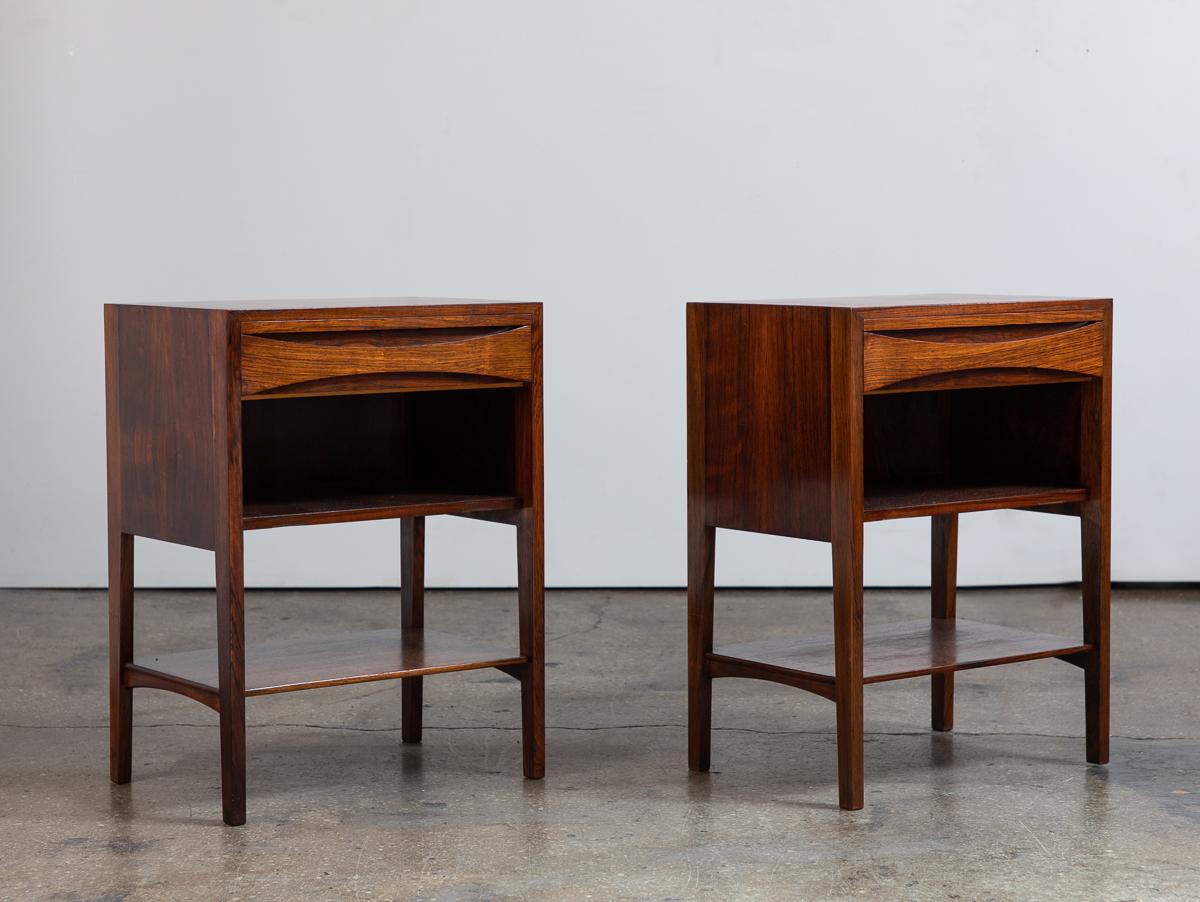 Pair of Danish Modern rosewood end tables, in the style of Arne Vodder. Drawer fronts feature an eye-catching carving with sweeping lines. Minimalist silhouette, with open shelving for additional storage and display. Hidden pull-out tray is a