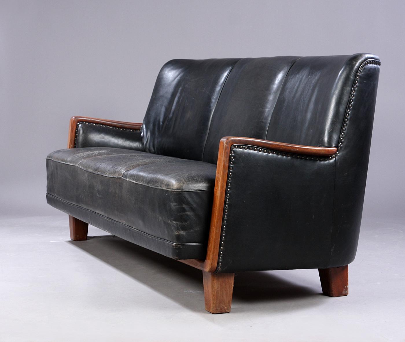 Danish cabinetmaker sofa, 1930-1940s. Comfortable, three-person. sofa, upholstered in partially stitched patinated black leather, mahogany armrests, lacquered wood and lacquered legs. Measures: H. 85/44, L. 200, D ca. 80 cm.