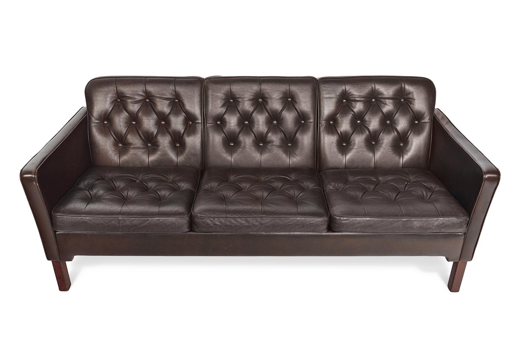 This stunning Danish modern chocolate brown patinated leather sofa offers rich detailing in a sleek, modern silhouette. This beautifully tapered frame holds six button tufted cushions. Sofa stands on stained beech legs. Upholstered on all sides. In