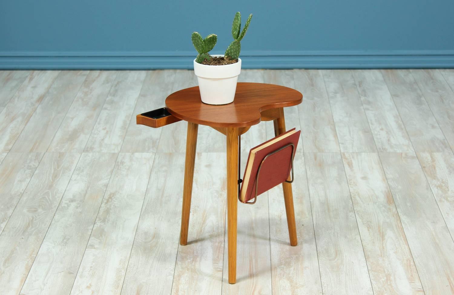 Peculiar side table manufactured by Møbelfabriken Gorm in Denmark circa 1950’s. This side table features a minimalist design with a teak table top and oak tapered legs. This exceptional model includes a hidden pull-out ashtray and a brass magazine
