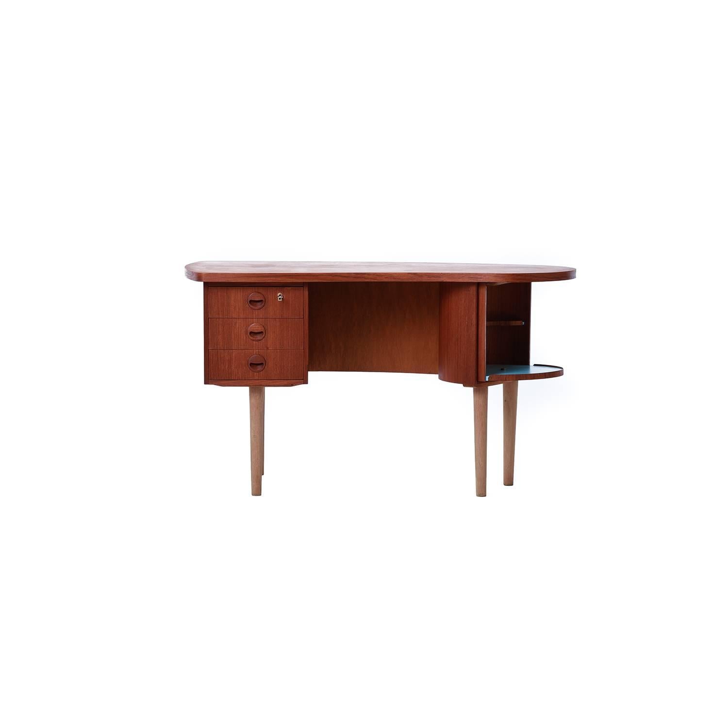 This petit kidney shaped teak desk with oak legs packs a punch with multiple storage options: three drawers, a lockable compartment, open back shelving, and a Lazy Susan bar with eggshell blue accent.
