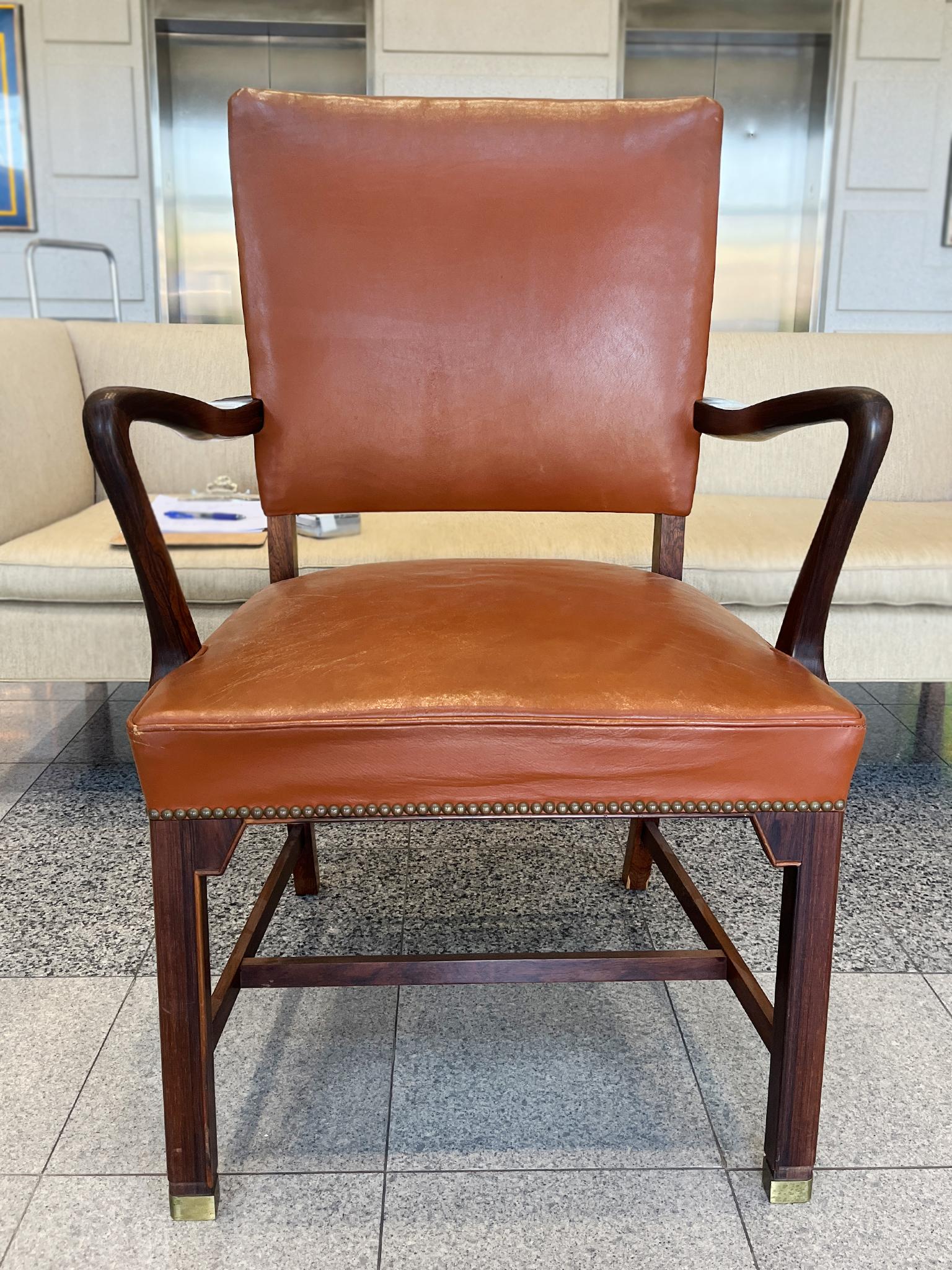 Realizzata negli anni '40, questa poltrona in stile Fritz Hansen è composta da una struttura in palissandro e da un rivestimento in pelle cognac con borchie in ottone. Un mix di forme arrotondate e lineari caratterizza il design complessivo della