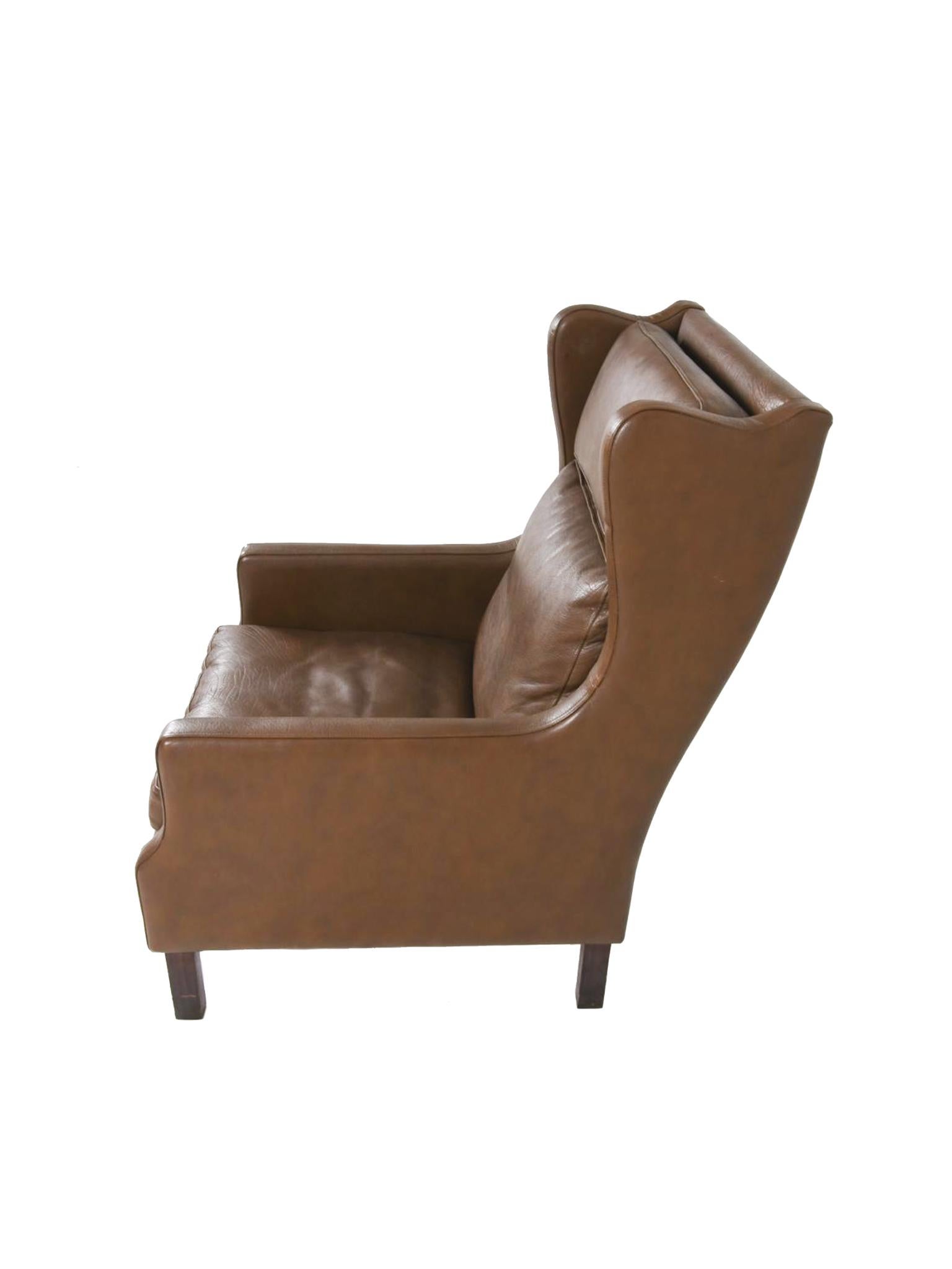 Ce fauteuil, qui s'inspire de la tradition des fauteuils à oreilles, est composé d'un revêtement en cuir marron et de pieds en bois de hêtre. Elle se distingue par ses lignes courbes et épurées et ses coins arrondis qui lui confèrent une allure