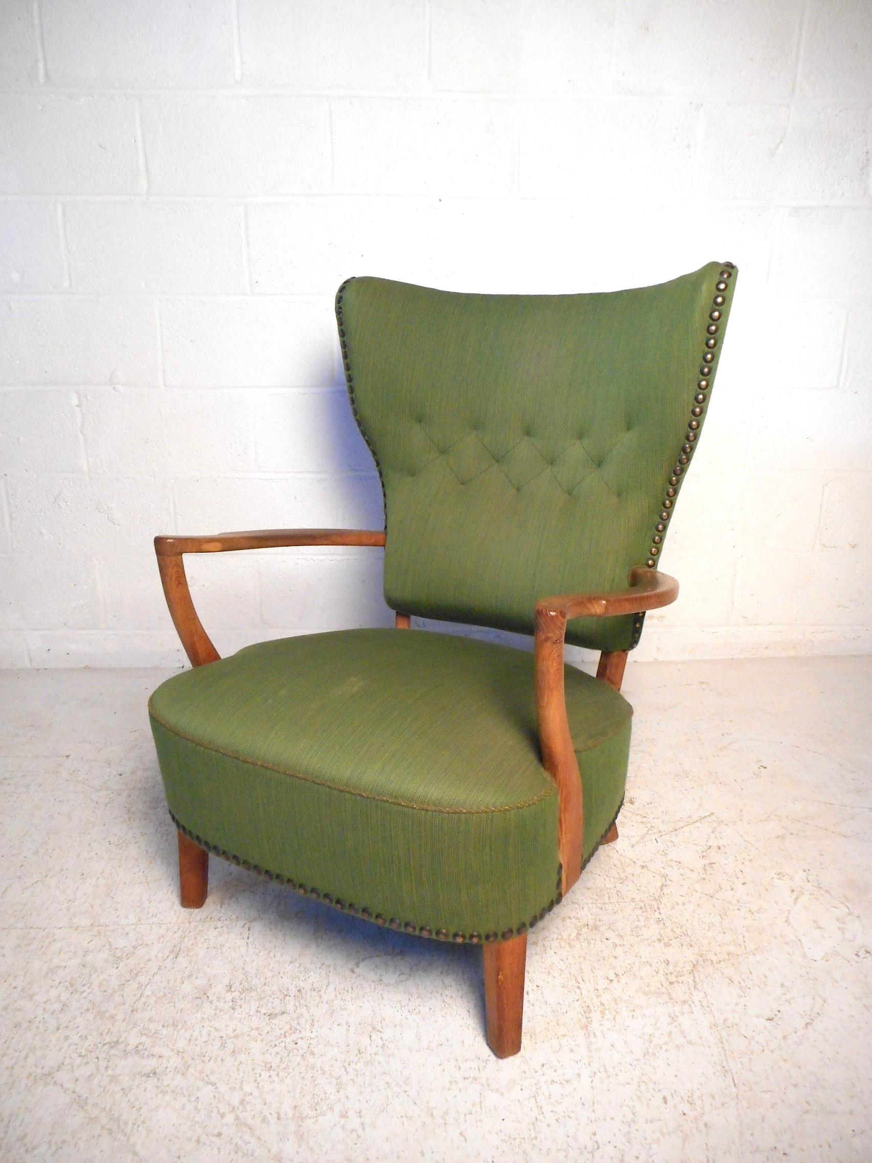 This impressive Danish modern chair features sculpted armrests, a tall winged back, and sturdy wooden legs. Covered with a vintage green upholstery, which has tufts on the backrest, and studs lining the contour of the chair. A great addition to any